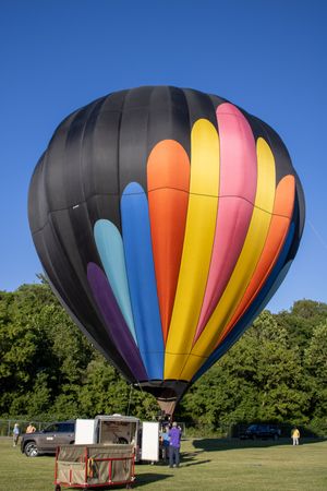 A vibrant striped hot air balloon stands inflated against a bright blue sky with trees in the background.
