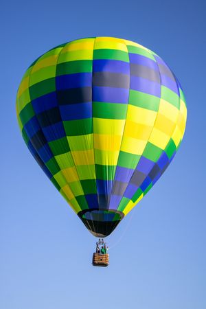 Hot air balloon with geometric pattern in green, yellow, blue and gray colors soaring in clear blue sky.