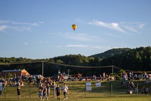 Hot air balloons float in the sky above a grassy field during a summer festival.