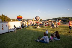 Panoramic view of hot air balloon festival with crowd gathered on grassy field at sunset.