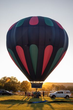 Black hot air balloon with red and green accents being prepared for launch at golden hour.