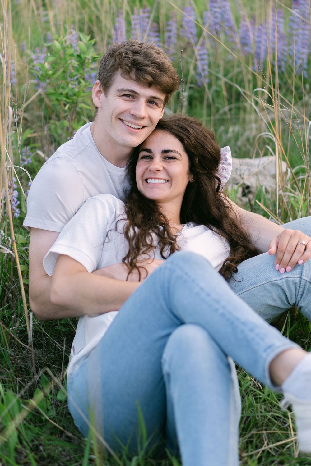 Quiet moment between a couple seated in a sunlit field, heads resting together with wildflowers and summer grass surrounding them