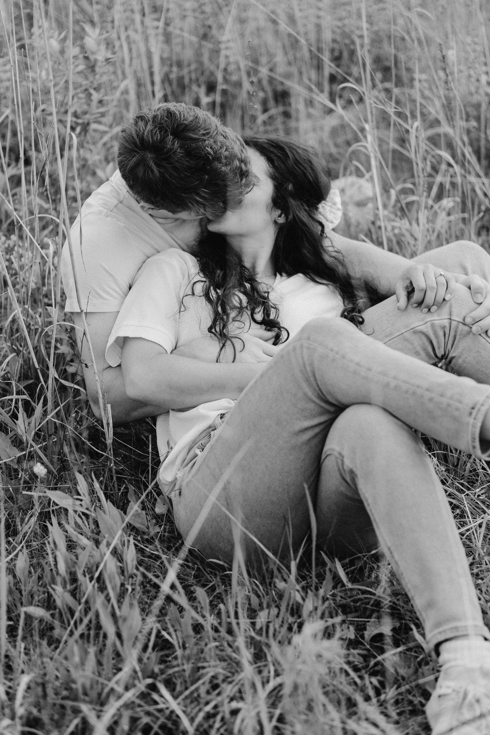Romantic engagement photo of couple relaxing in a grassy clearing, wrapped in each other’s arms with soft sunset light behind them in Hudson