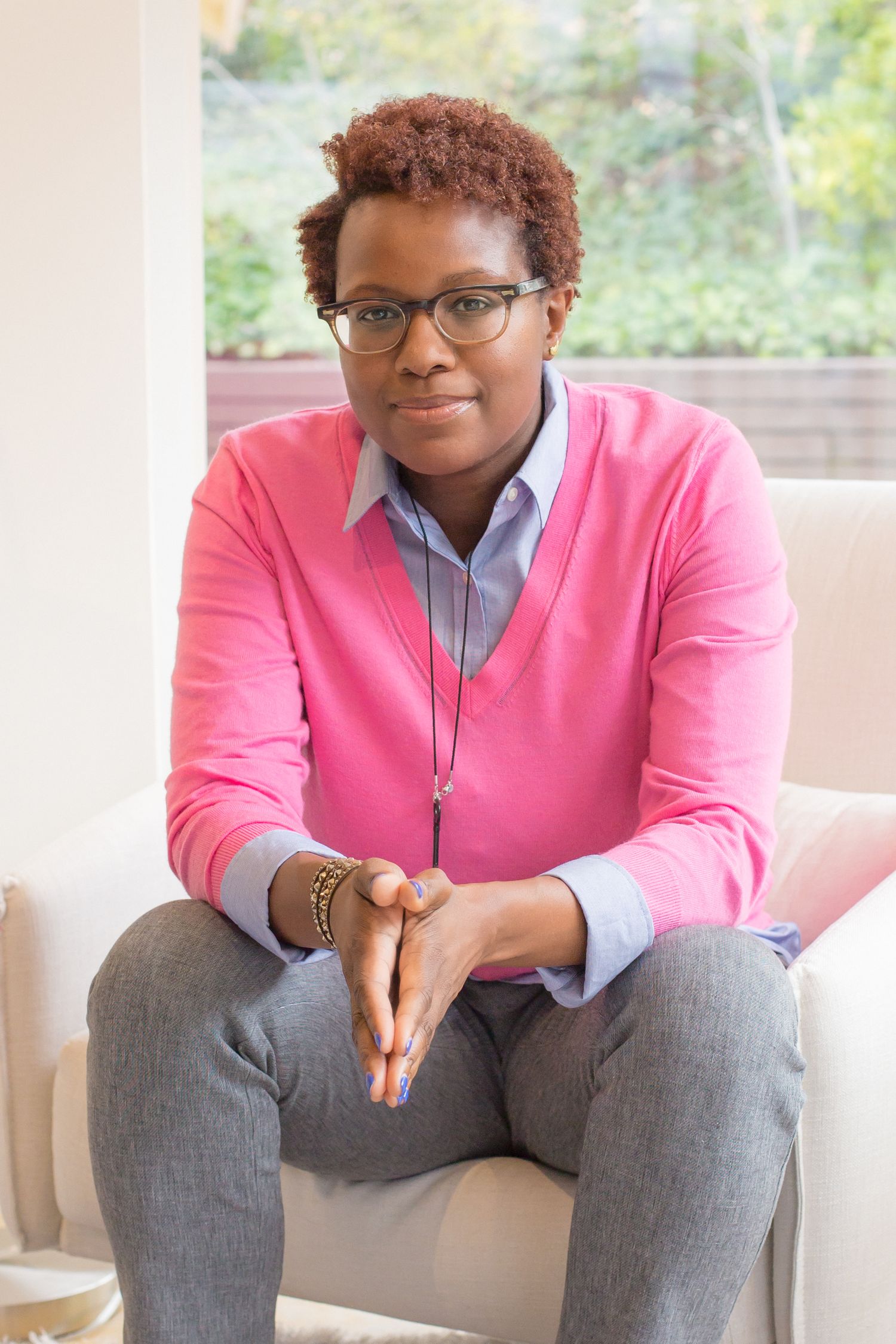 Professional seated portrait in bright pink v-neck sweater and gray button-down shirt with glasses and short curly hair.