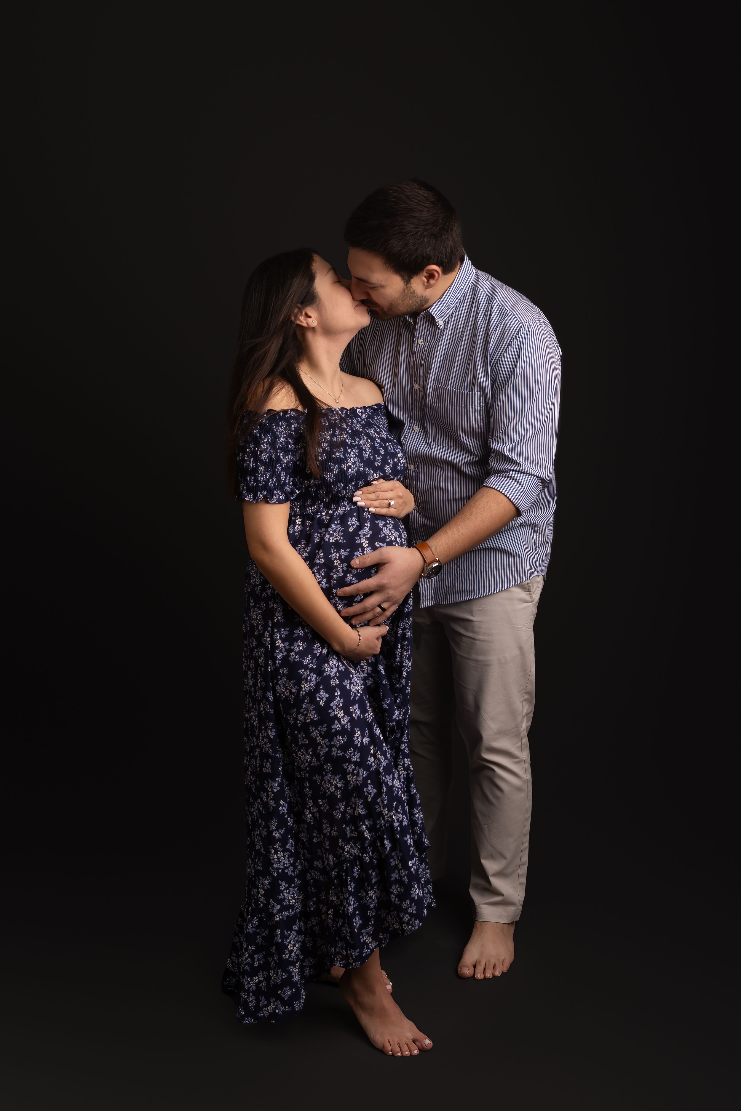 A couple embraces intimately in a dimly lit studio setting wearing a navy floral dress and gray button-up shirt.