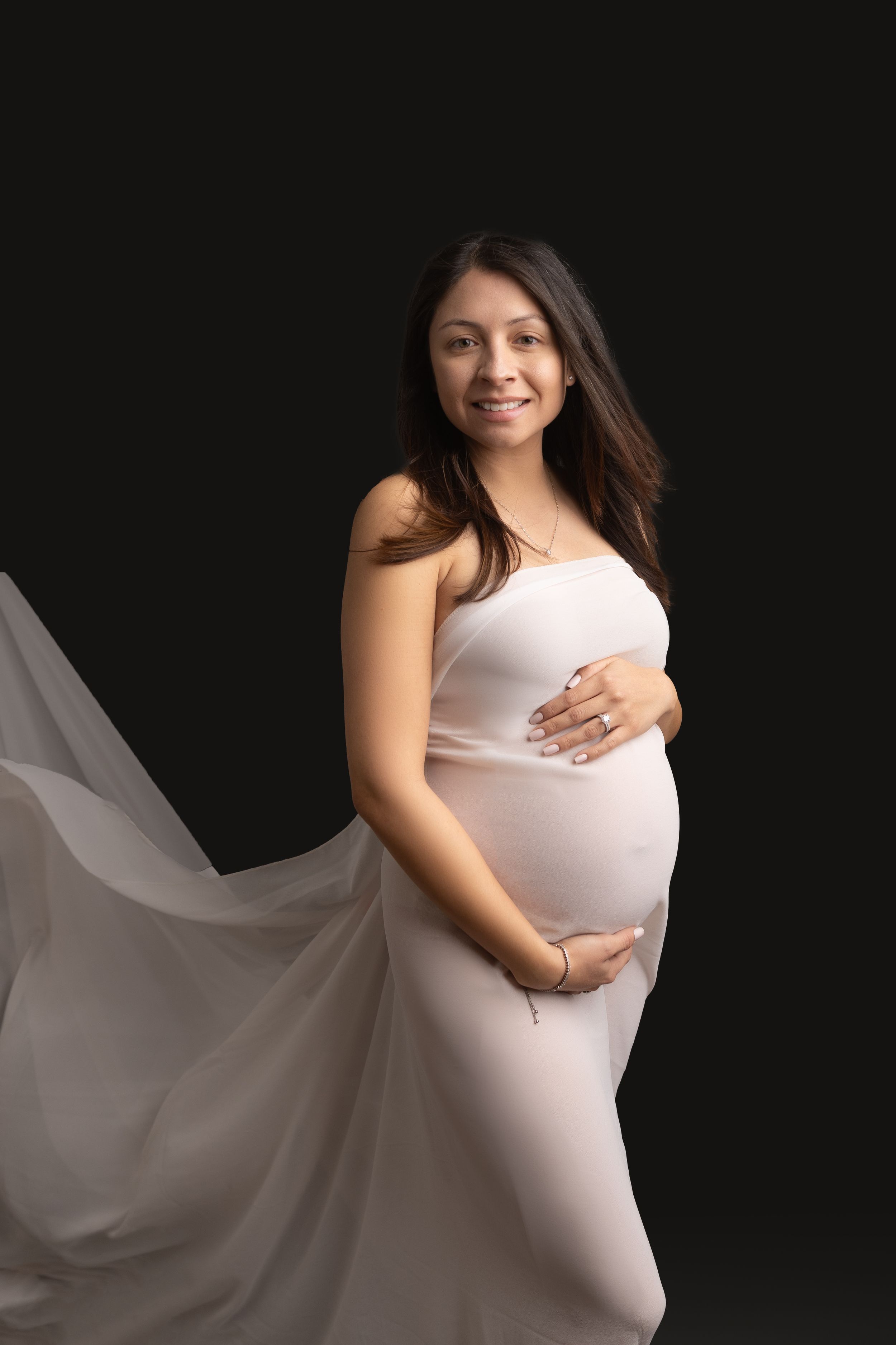 Maternity portrait featuring an elegant white gown with flowing fabric against a dark background.