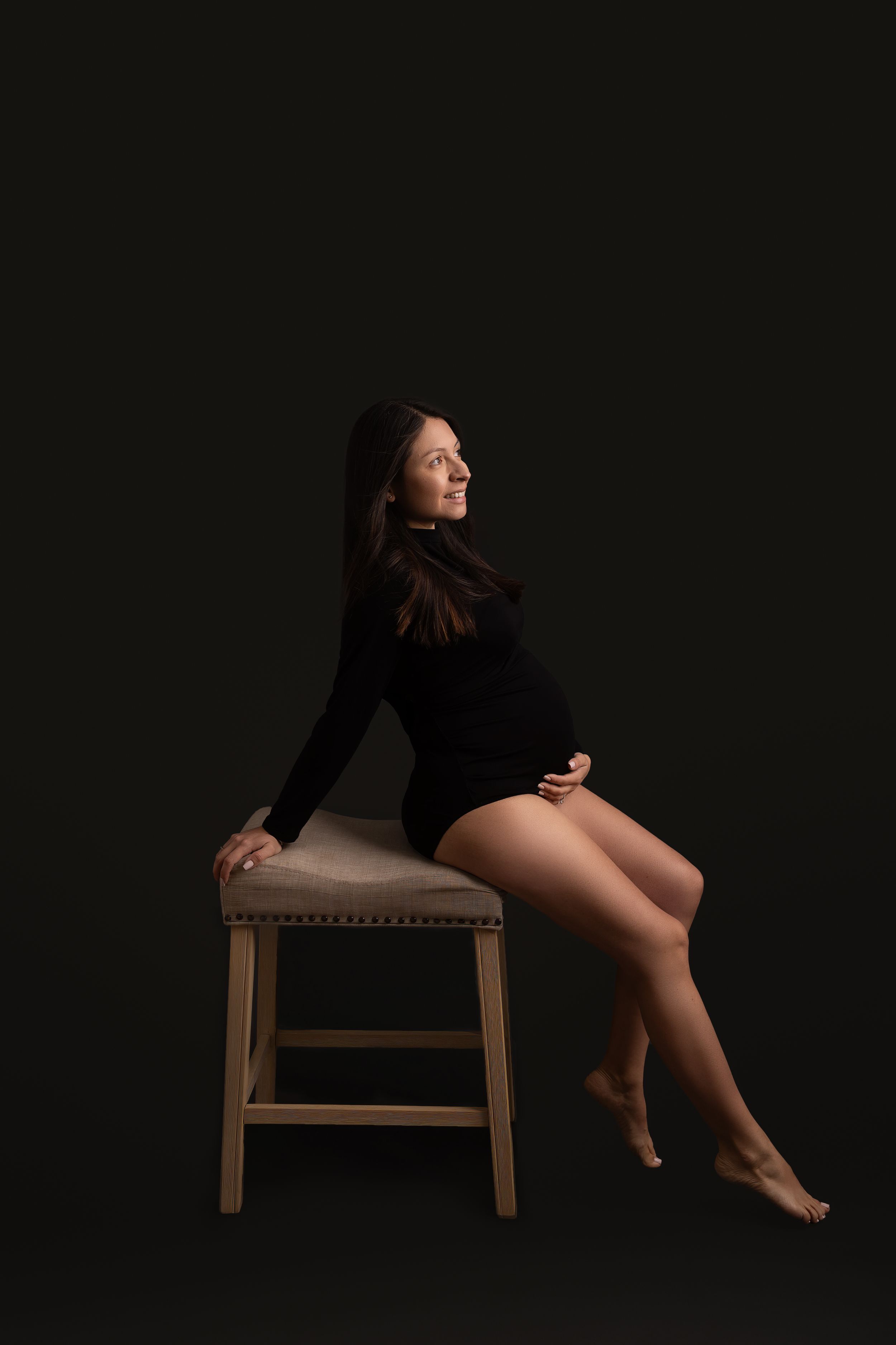 Dark artistic maternity photo of someone posing gracefully in black bodysuit on wooden stool against black background.
