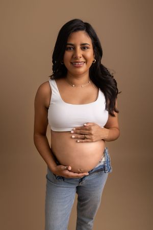 Expecting mother in white crop top and jeans poses against brown backdrop for maternity photo.