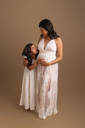 A mother and daughter share a tender moment together wearing matching white dresses against a neutral brown backdrop.