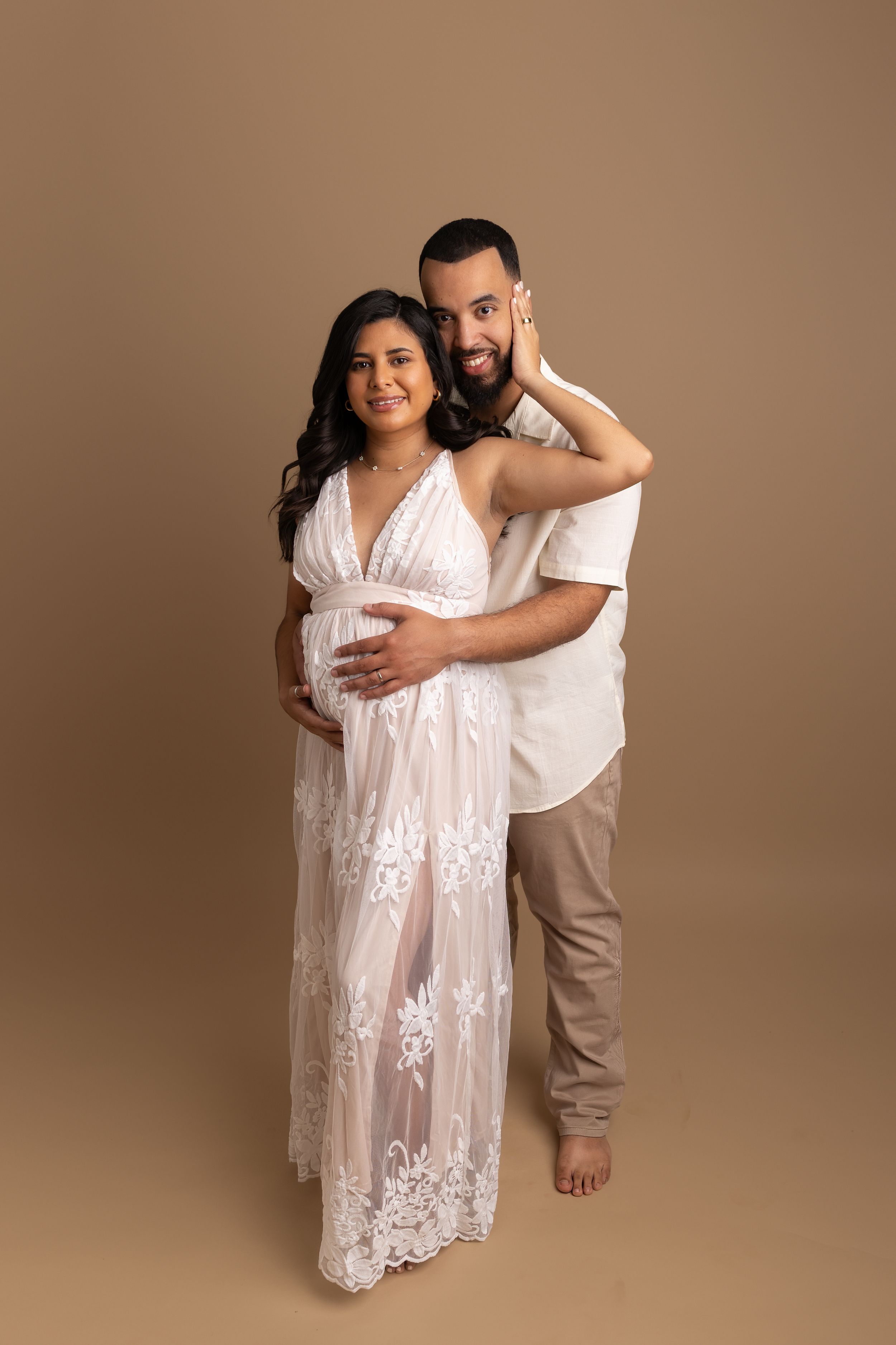 Couple in white outfits embrace during intimate maternity photoshoot against warm brown background.
