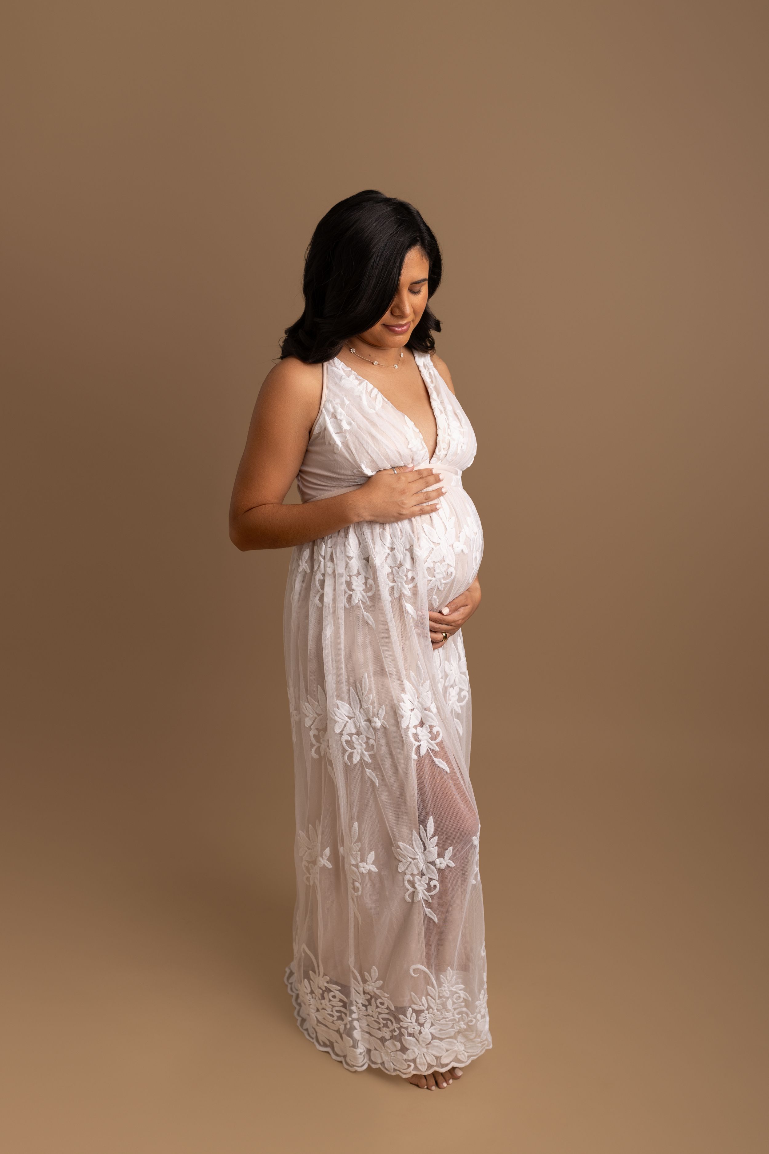 Maternity photo session showing person in flowing white lace dress against neutral beige background.