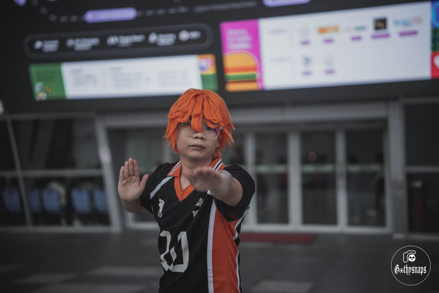 Cosplayer in a black and orange volleyball uniform waves at the camera during a convention.