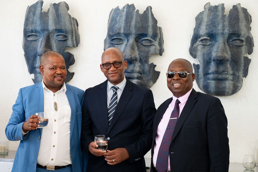 Three men in business attire stand in front of large blue-tinted sculptural faces mounted on a light colored wall.