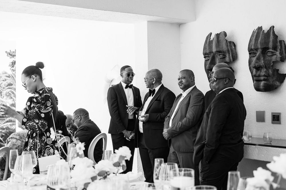 A formal black and white photograph shows people gathered at an elegant dining table with tribal masks visible on the wall.
