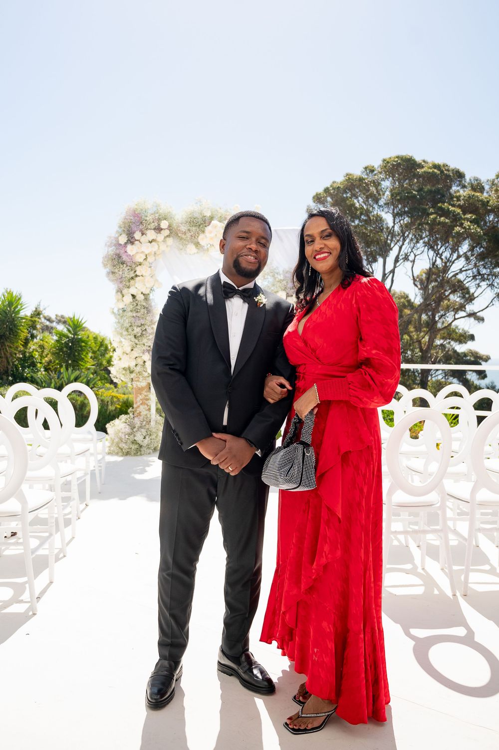 A couple stands together outdoors at a wedding venue, with one person wearing a black tuxedo and another in a red dress.