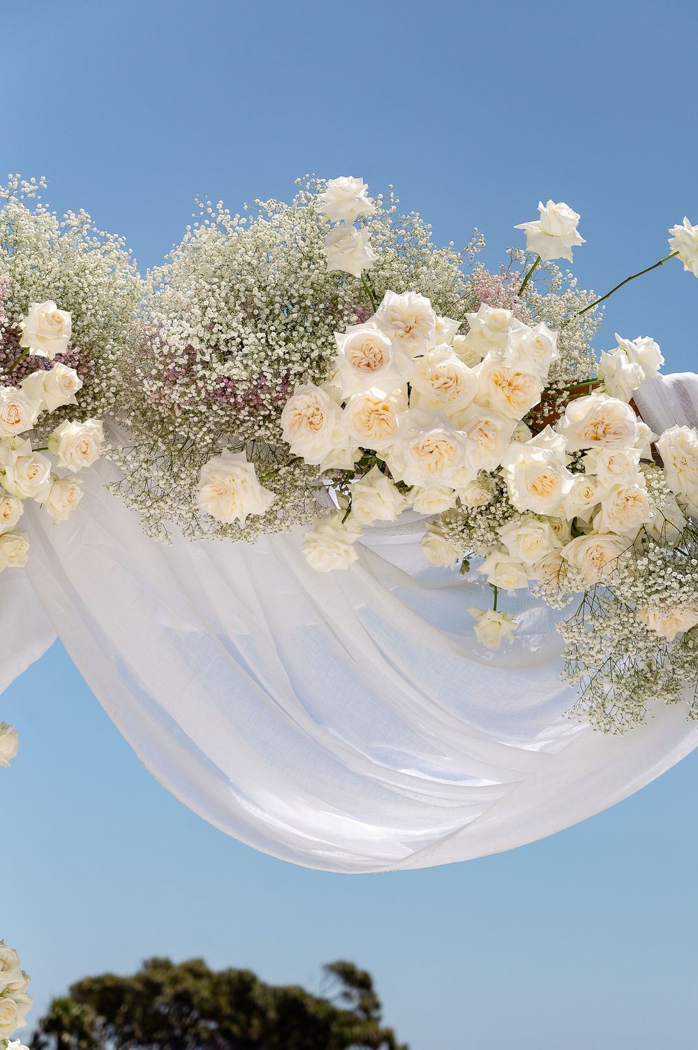 White fabric draping adorned with cream roses and delicate baby's breath flowers under blue skies.