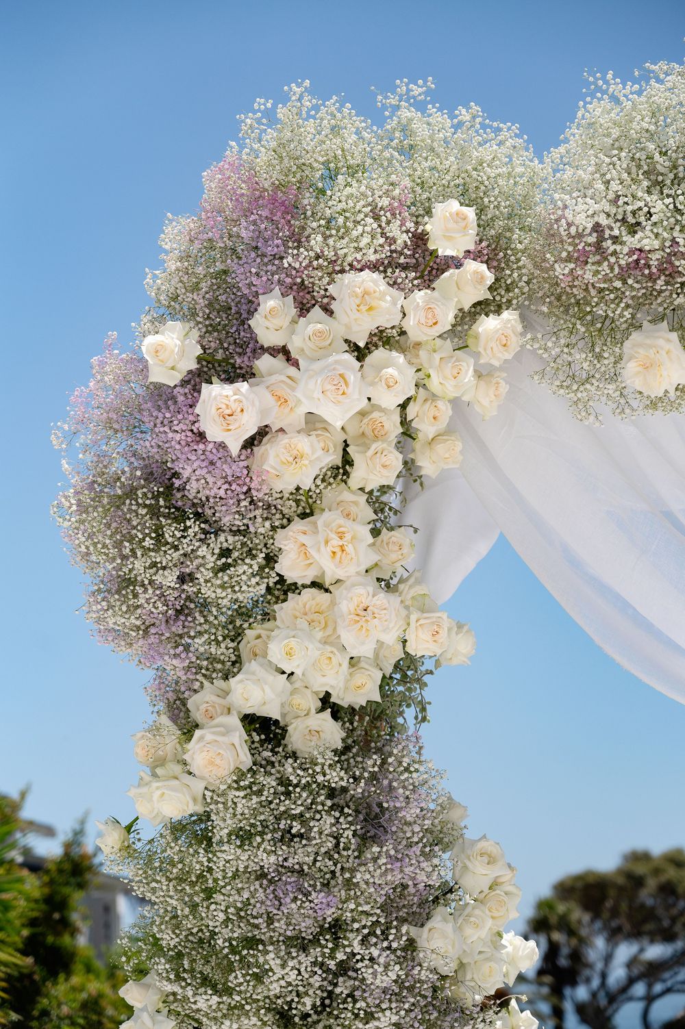 Elegant wedding arch decorated with white roses, baby's breath, and purple flowers against a bright blue sky.