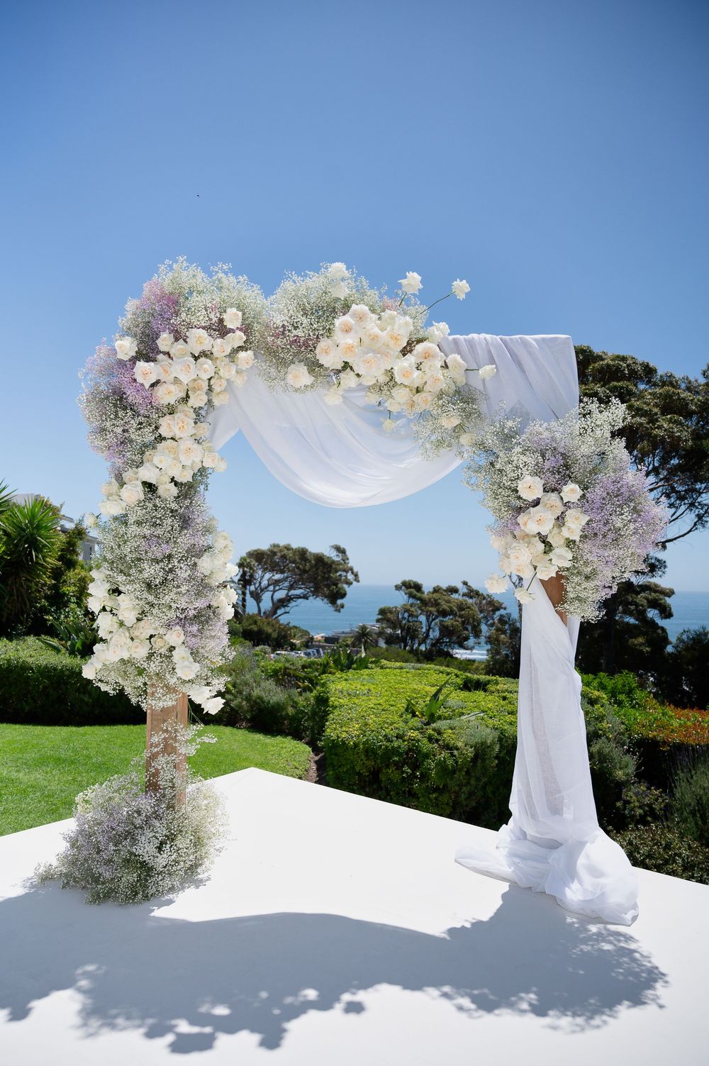 A white floral wedding arch decorated with roses and draped fabric stands on a grassy hilltop against a blue sky.