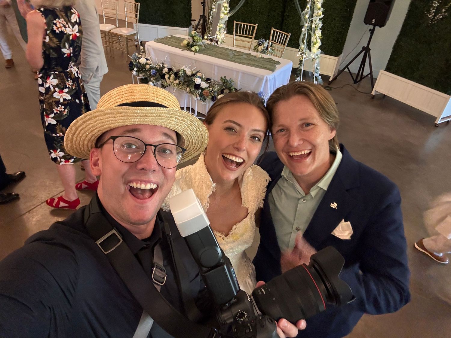 Three people take a festive selfie at what appears to be a wedding reception event with outdoor lighting.