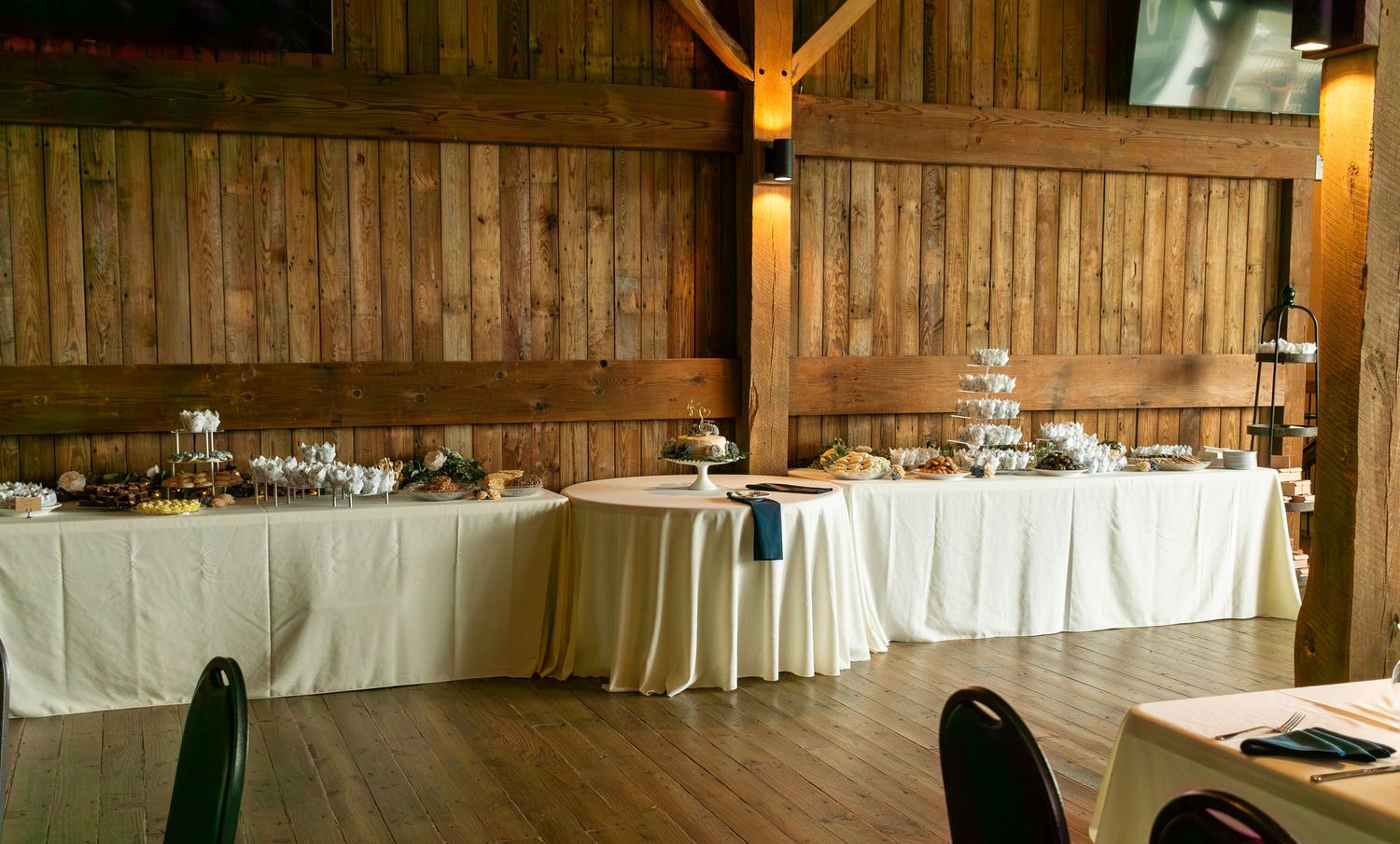 Long buffet table with white linens and serving dishes set up in a rustic wooden barn venue for catering service.