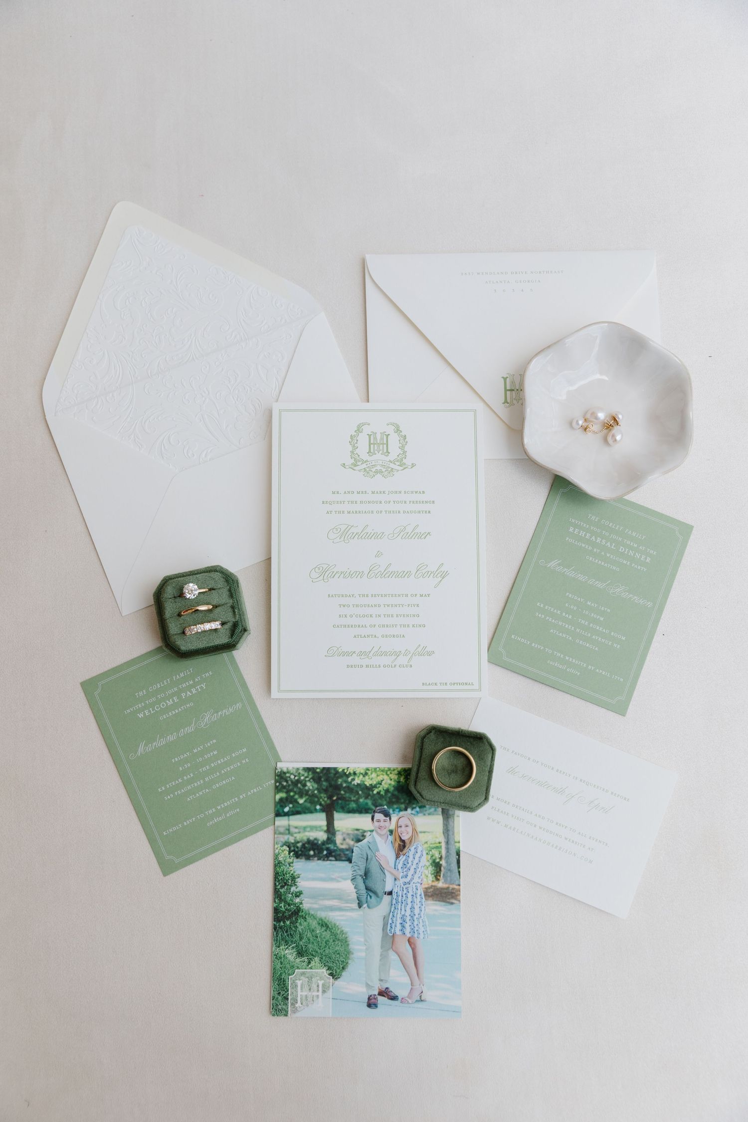 Wedding invitation suite in white and sage green with rings and small decorative dish on light background.