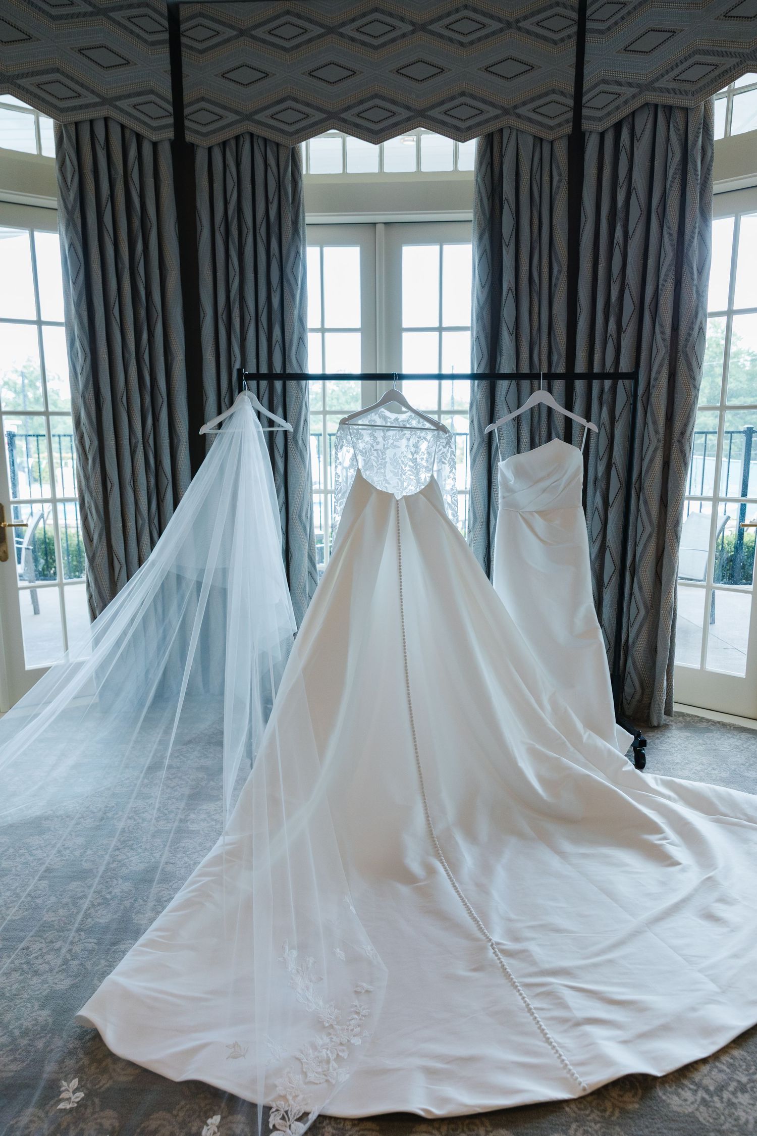 A sequence of wedding gowns displayed in a bright sunlit room with large windows and gray curtains.