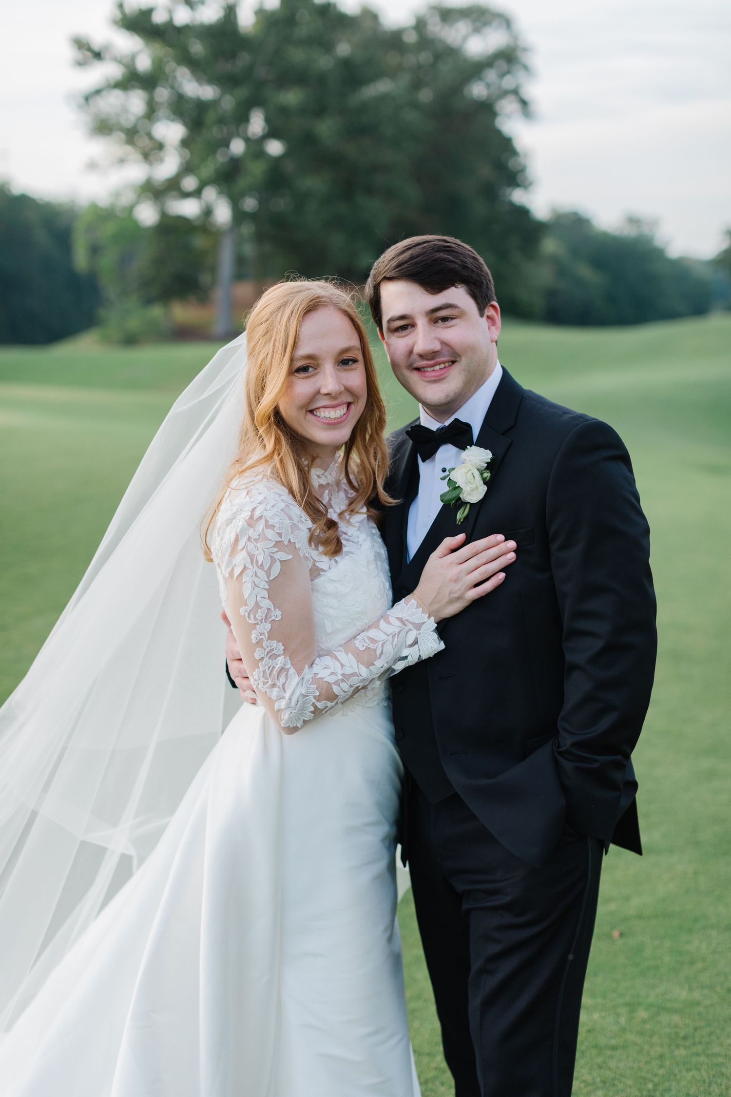 A wedding couple stands together on a golf course with lush green trees in the background.
