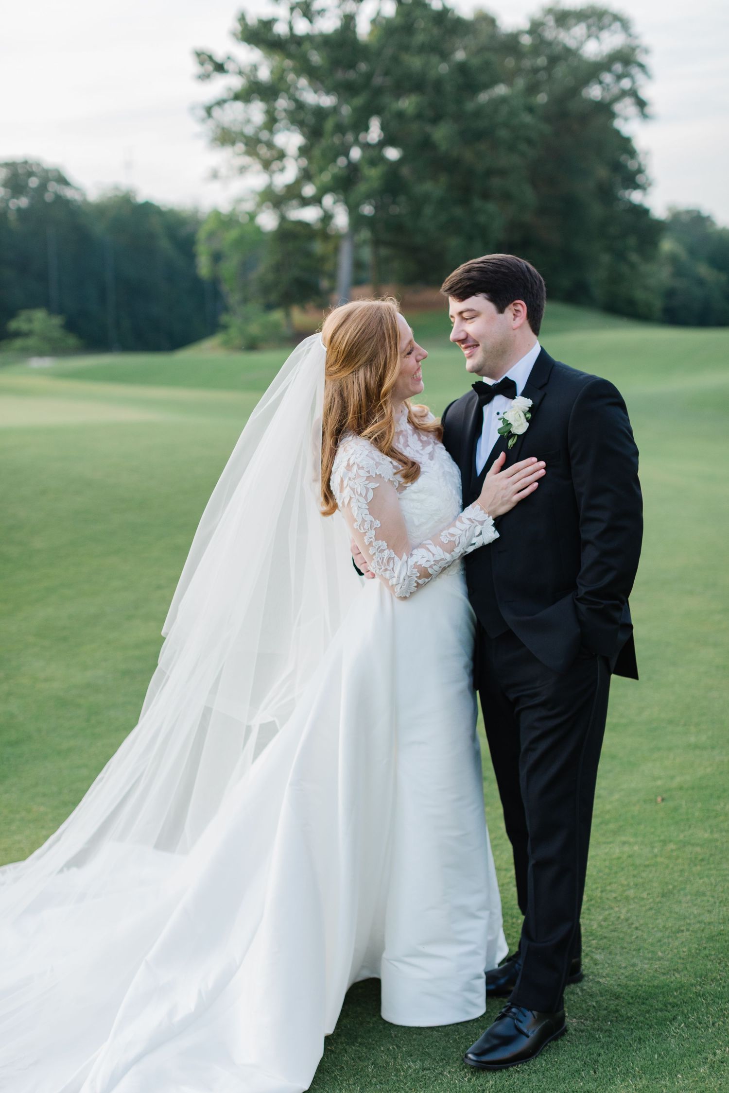 A romantic wedding couple embraces on a lush green golf course with a flowing white veil in the breeze.