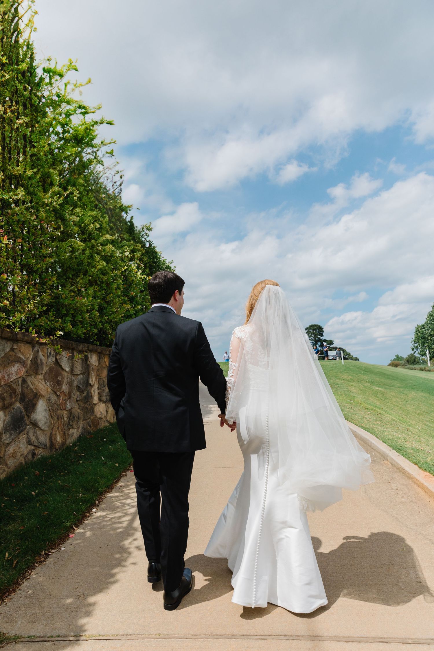 A couple in wedding attire walk hand in hand down a stone-lined path under blue skies on their special day.