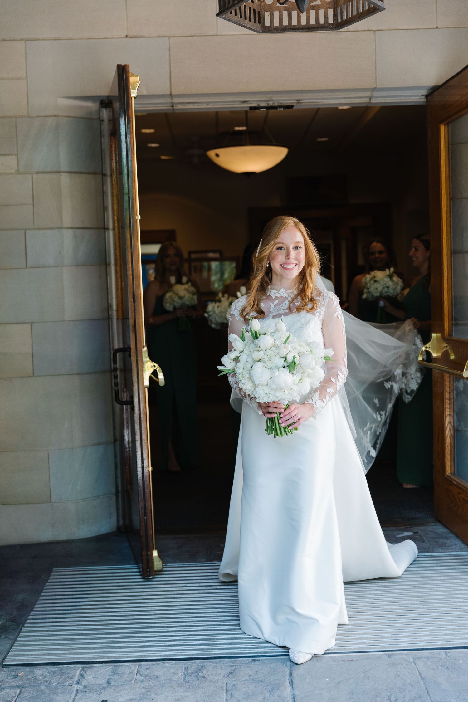 A bride in a long-sleeved white wedding gown holds a white floral bouquet while standing in a doorway.