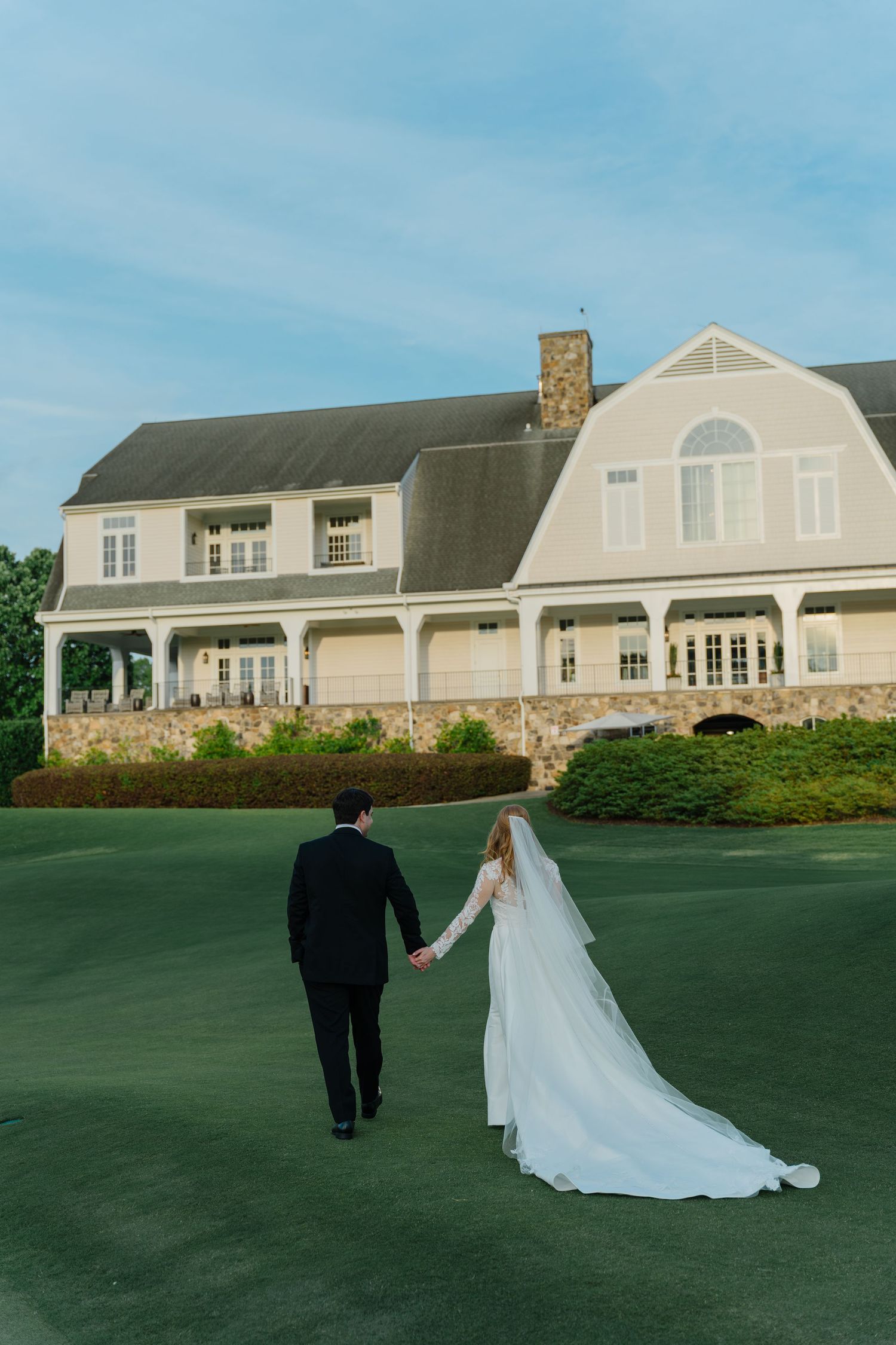 A couple in wedding attire walks across a manicured green lawn with a large beige luxury hamptons in the background.
