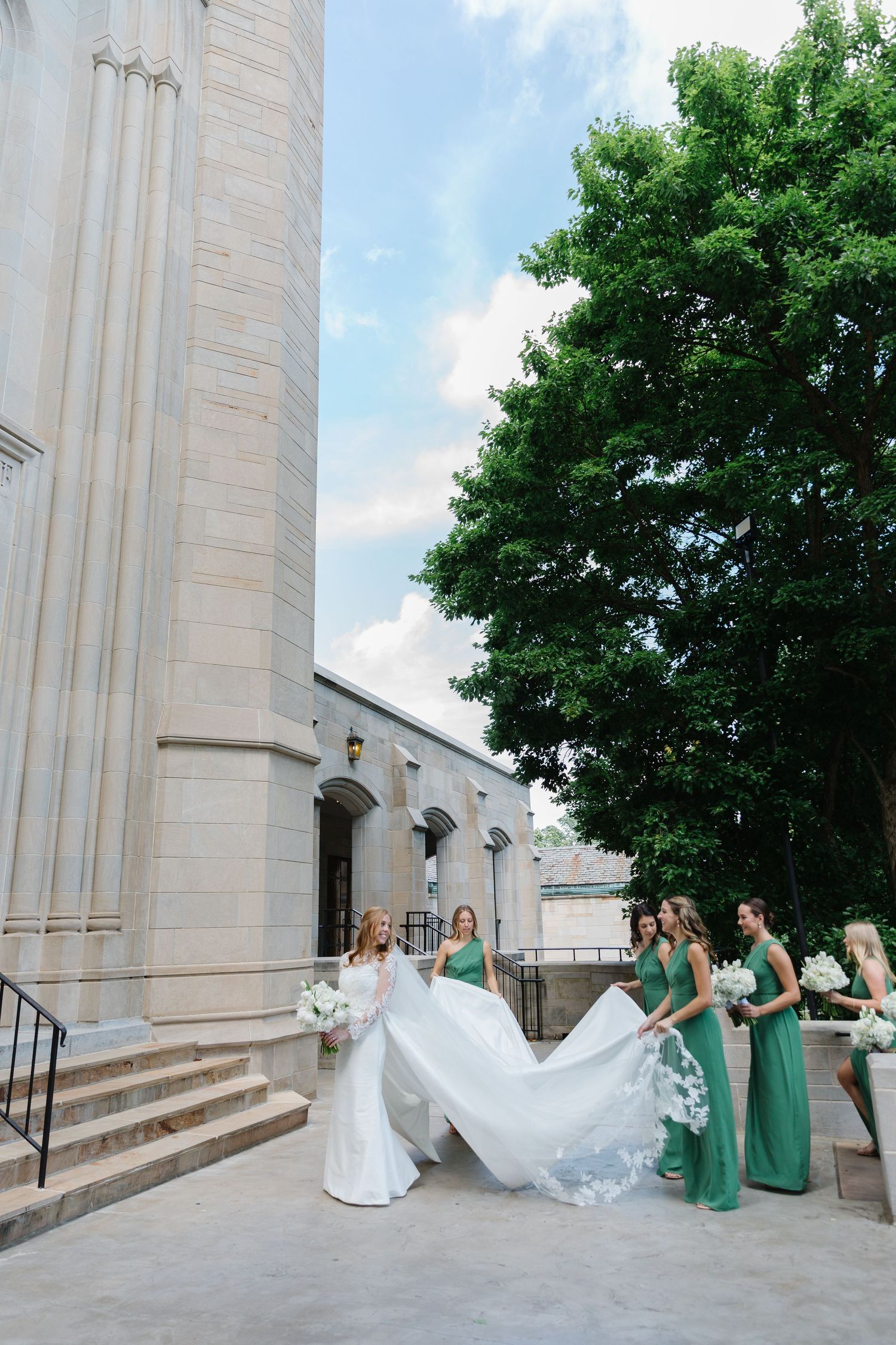A wedding party in emerald green dresses helps arrange a flowing white bridal train on church steps.