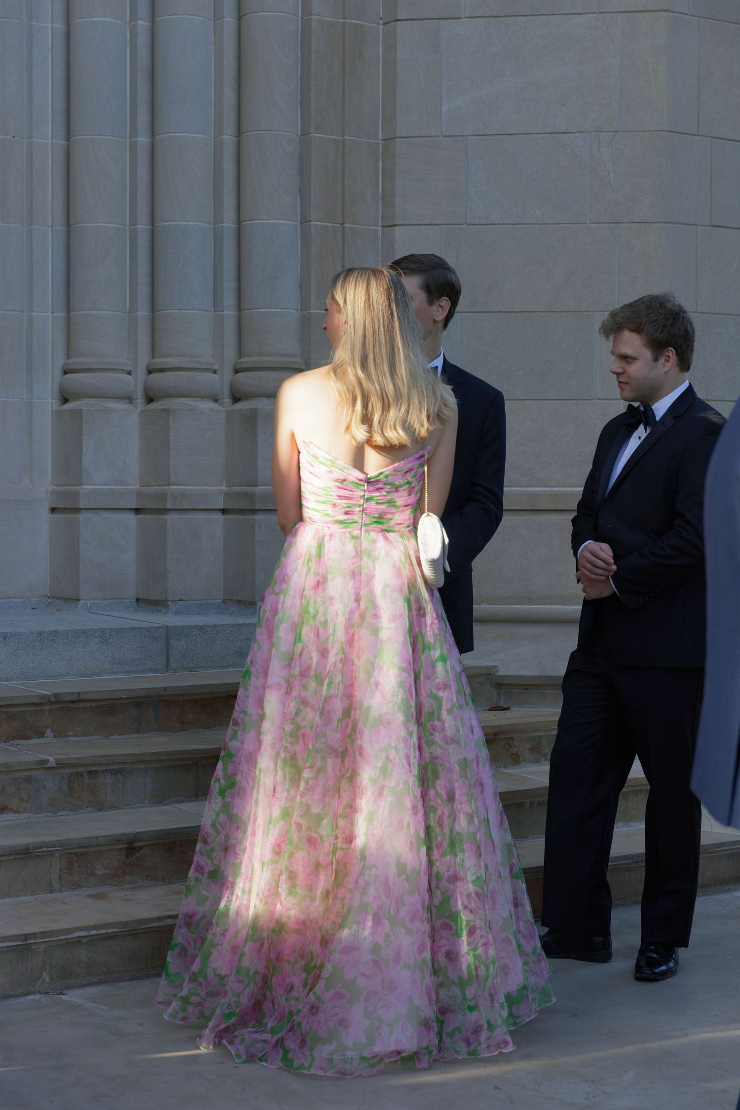 A couple in formal attire stands near stone columns, with the person wearing a floral print ballgown in pastel colors.