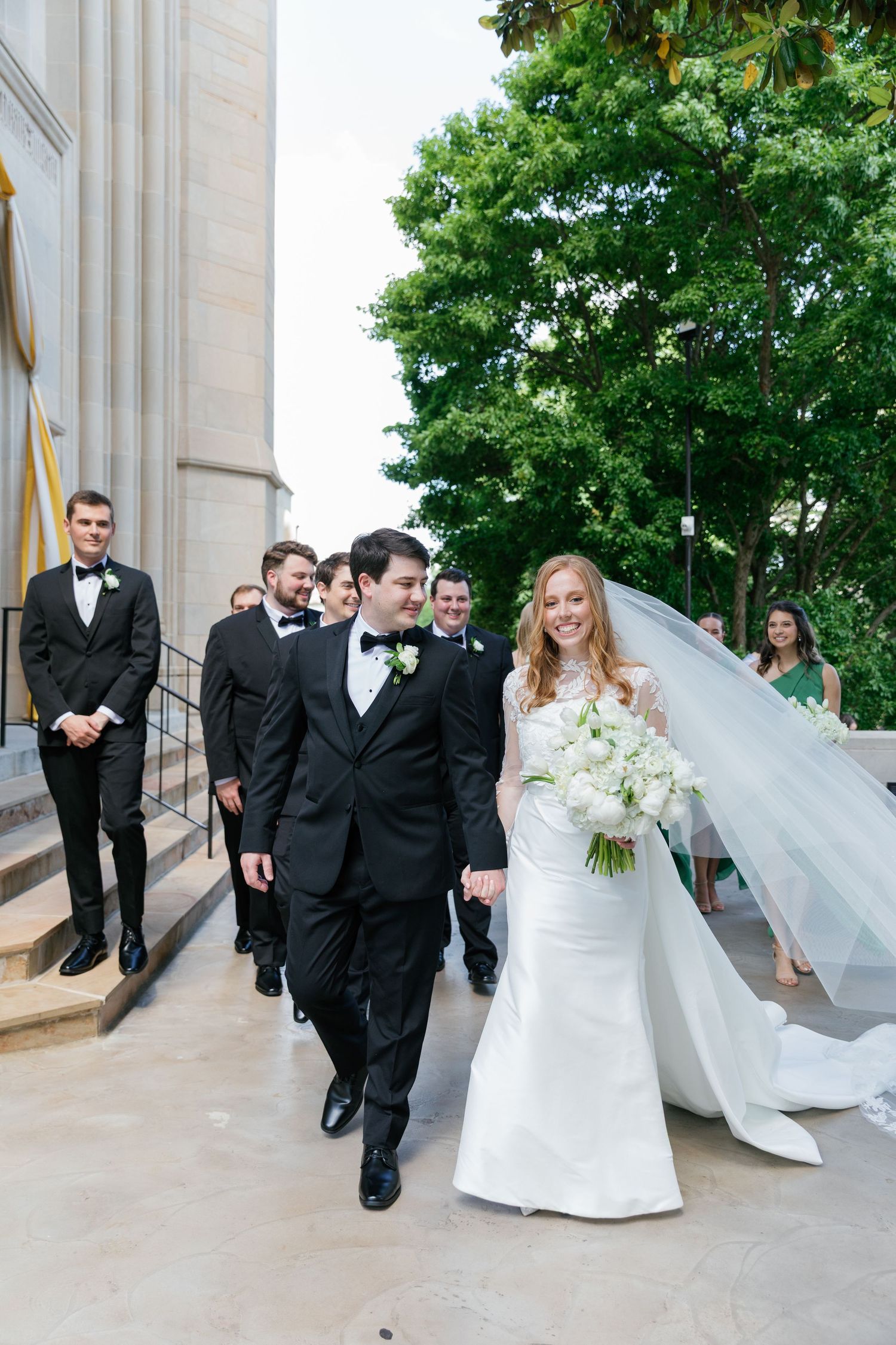 Wedding party poses on elegant stone steps with flowing white veil catching the breeze in outdoor ceremony setting.
