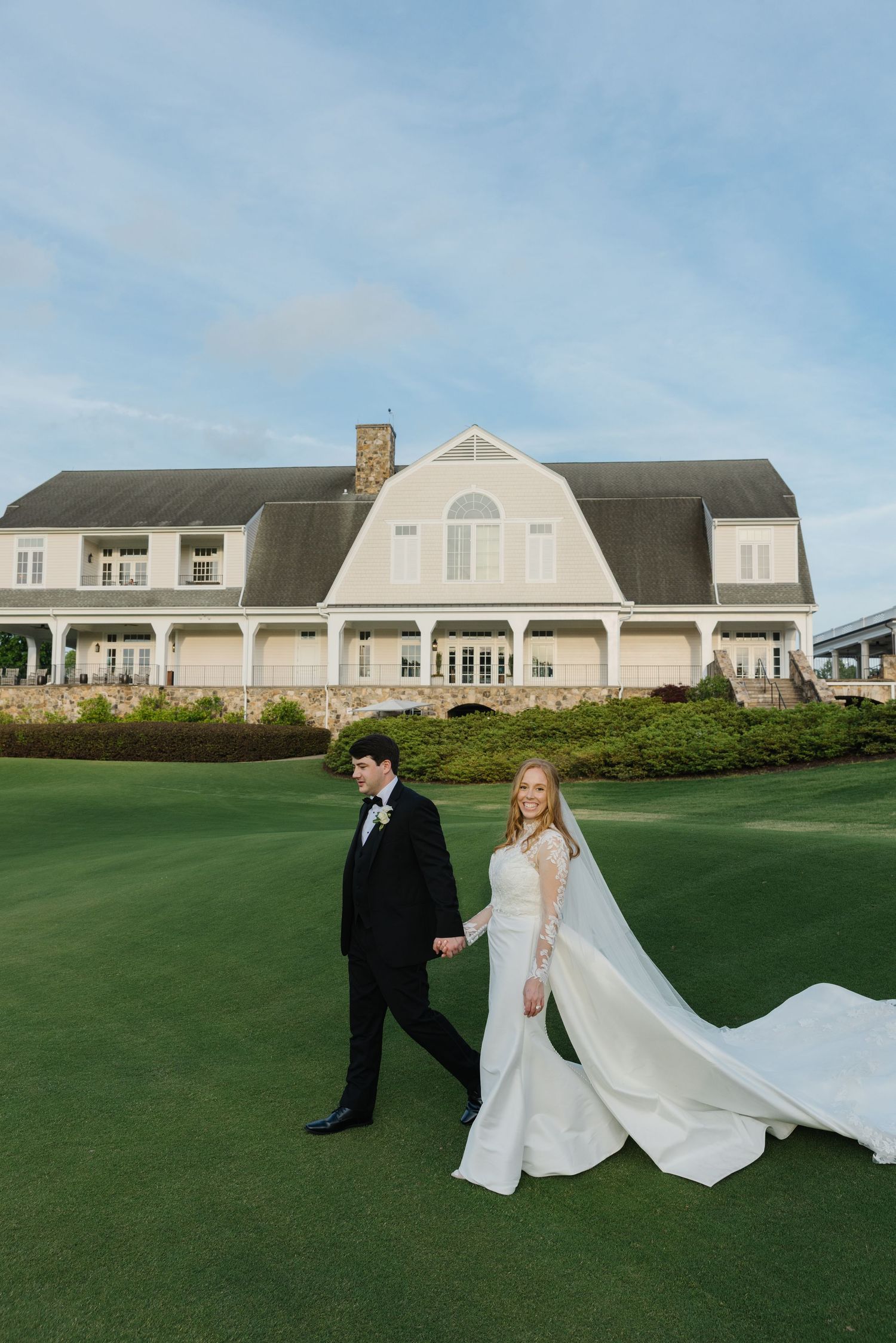 A couple in wedding attire poses on a manicured lawn with a large Hamptons clubhouse building in the background.
