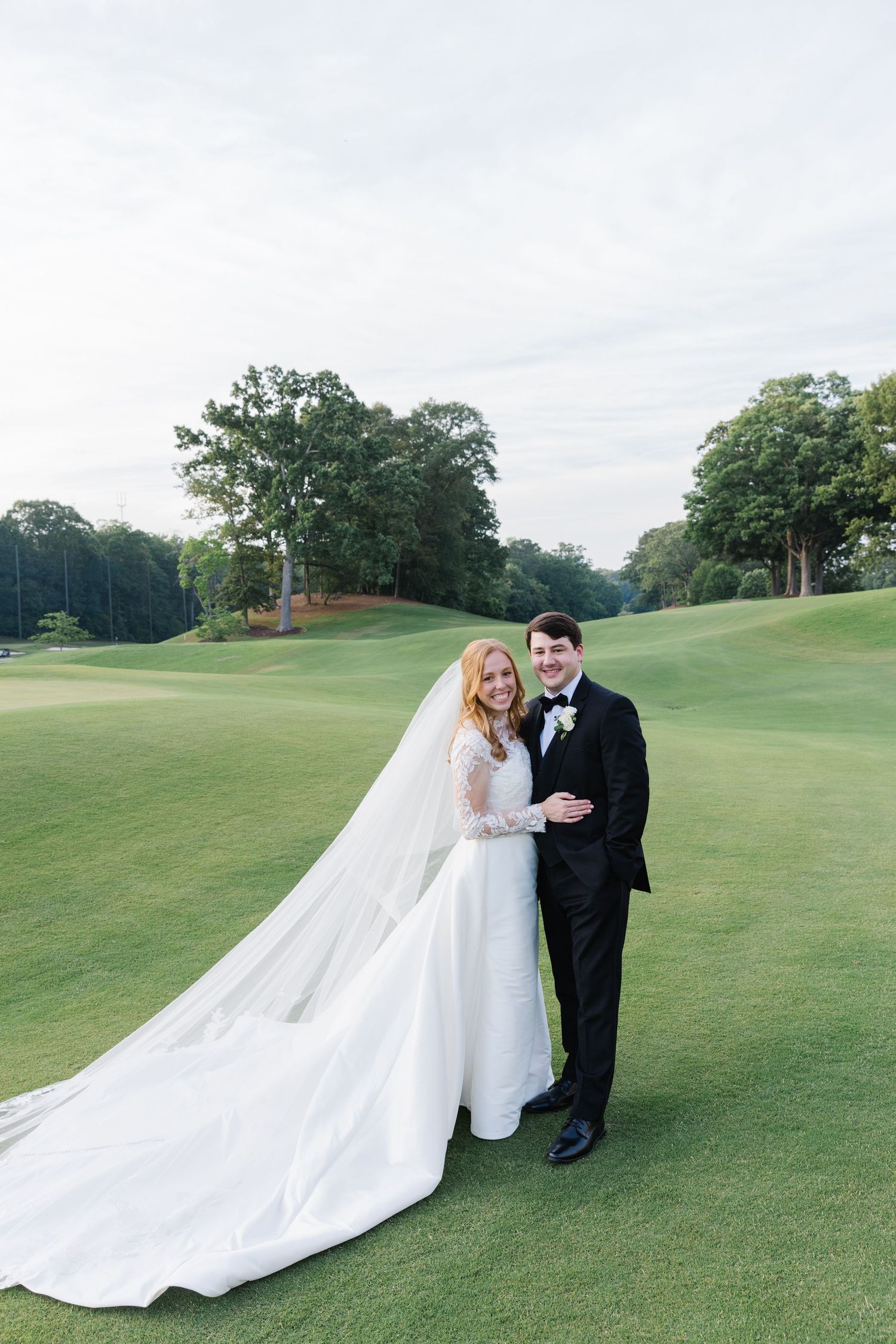 A couple poses together on a lush green golf course with a flowing white wedding dress trailing behind them.
