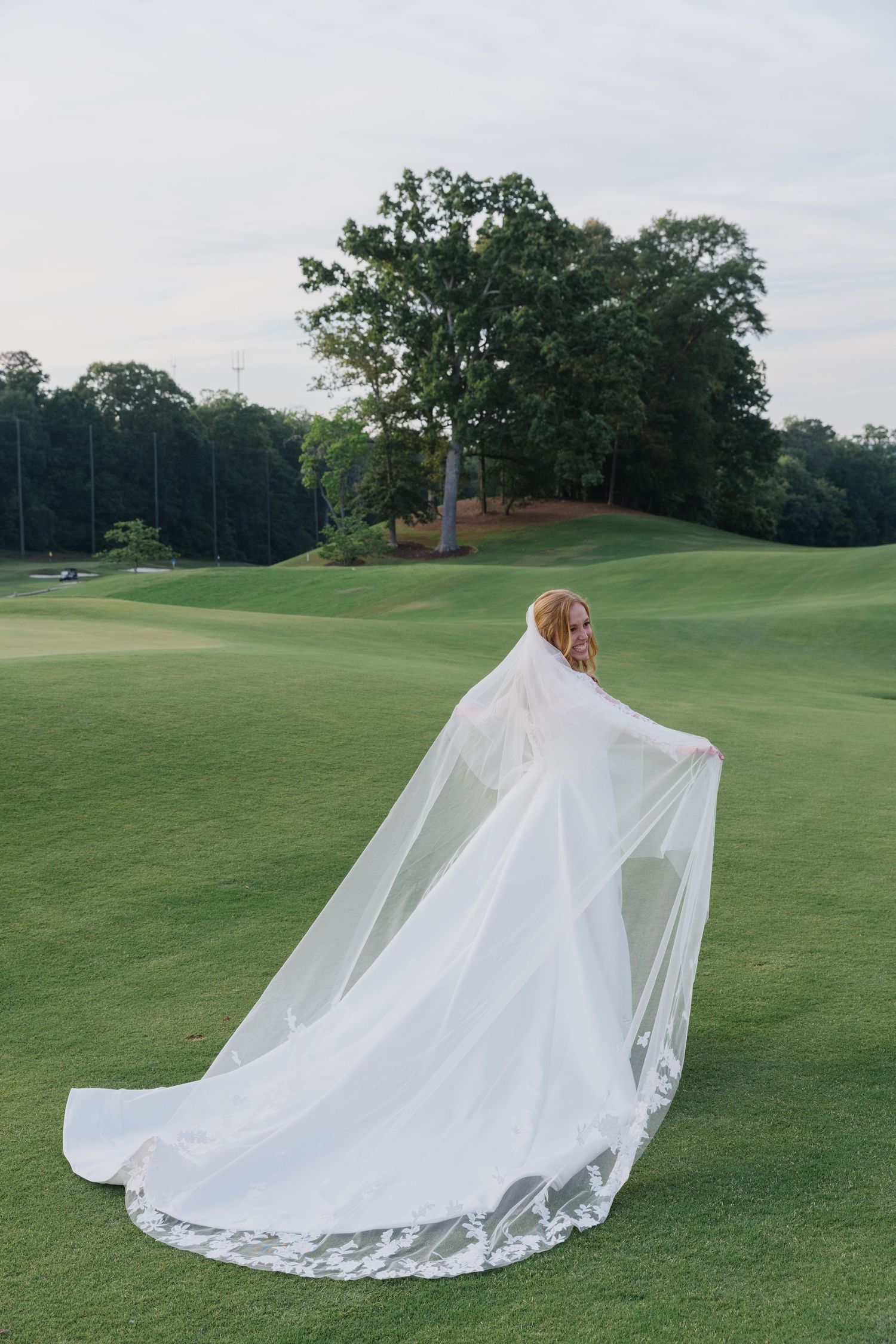 A flowing white wedding dress with lace trim billows dramatically across a lush green golf course.