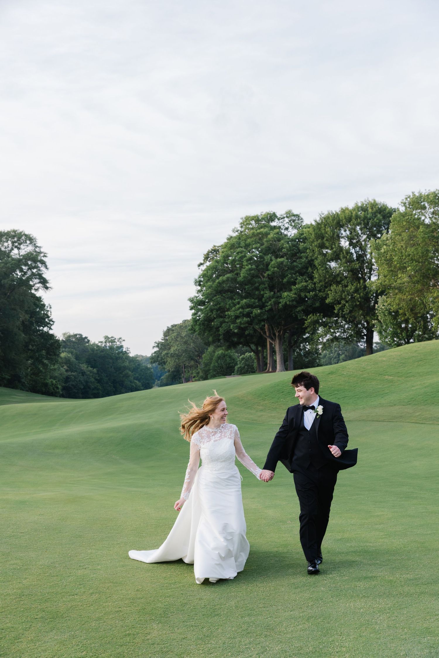 A couple in wedding attire runs playfully across a lush green hillside on their special day.