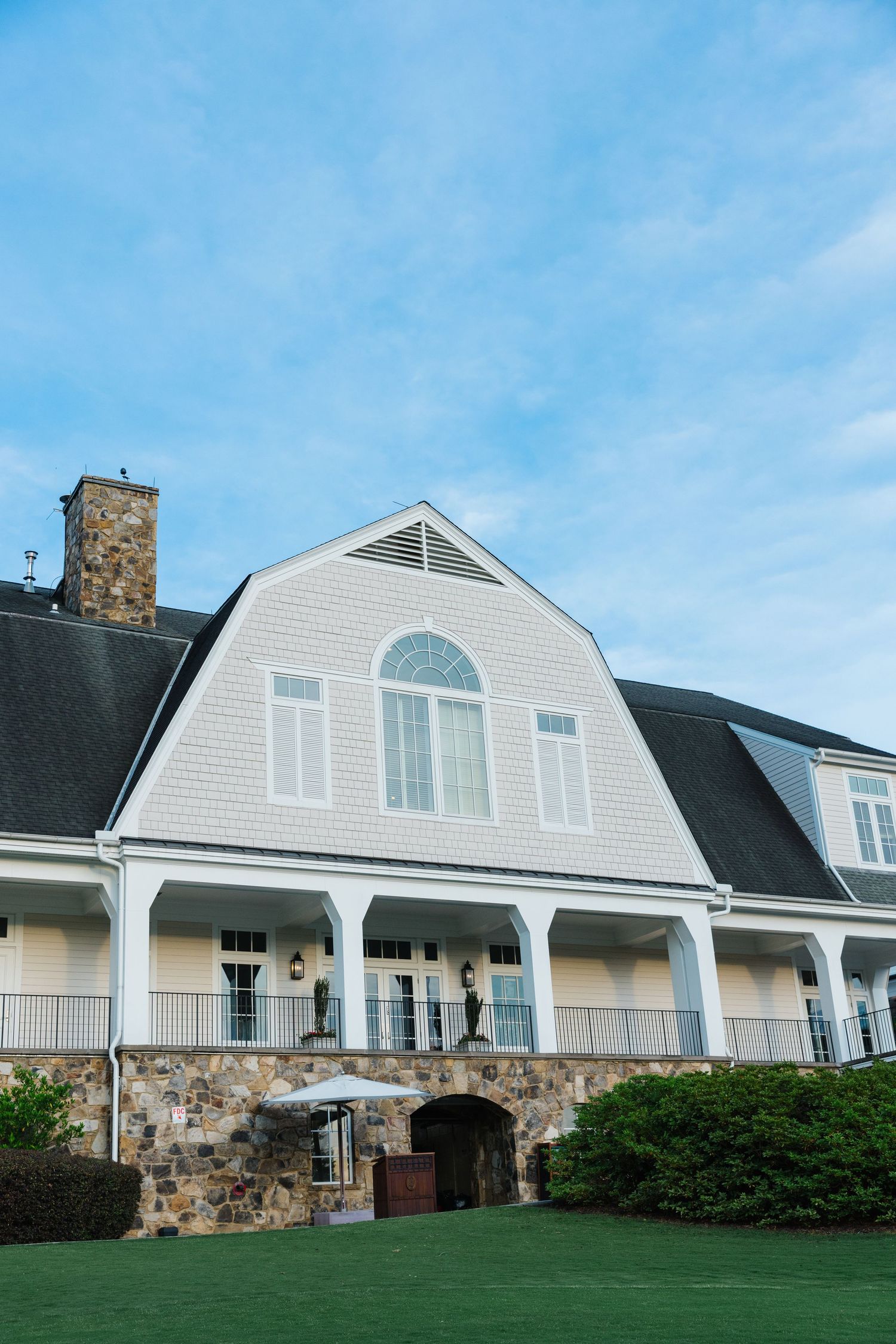 Panoramic view of a white gambrel-style mansion with stone accents and arched entryway set against a bright blue sky.