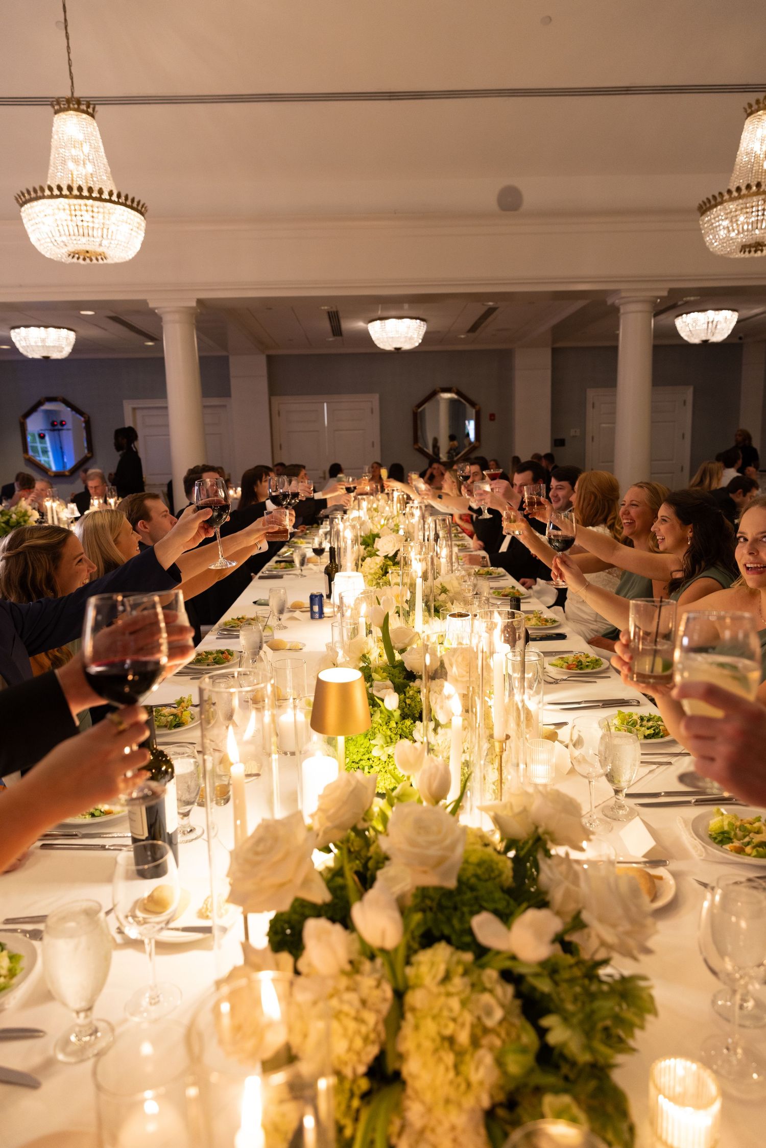Elegant wedding reception table with white floral centerpieces and glowing candles as guests raise their glasses in celebration.
