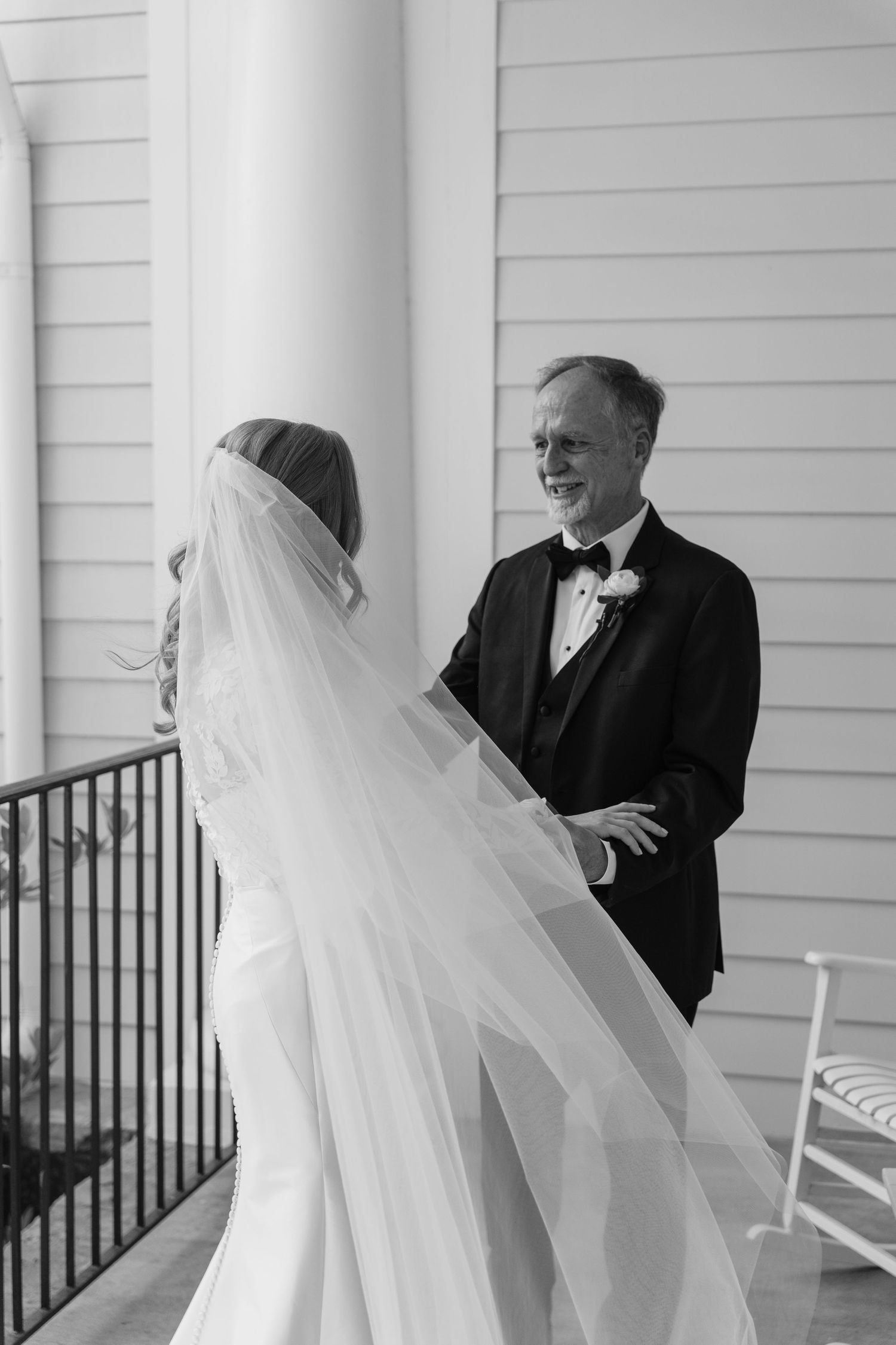 A black and white photo of a bride in a flowing veil with her companion on a porch with white columns and railings.