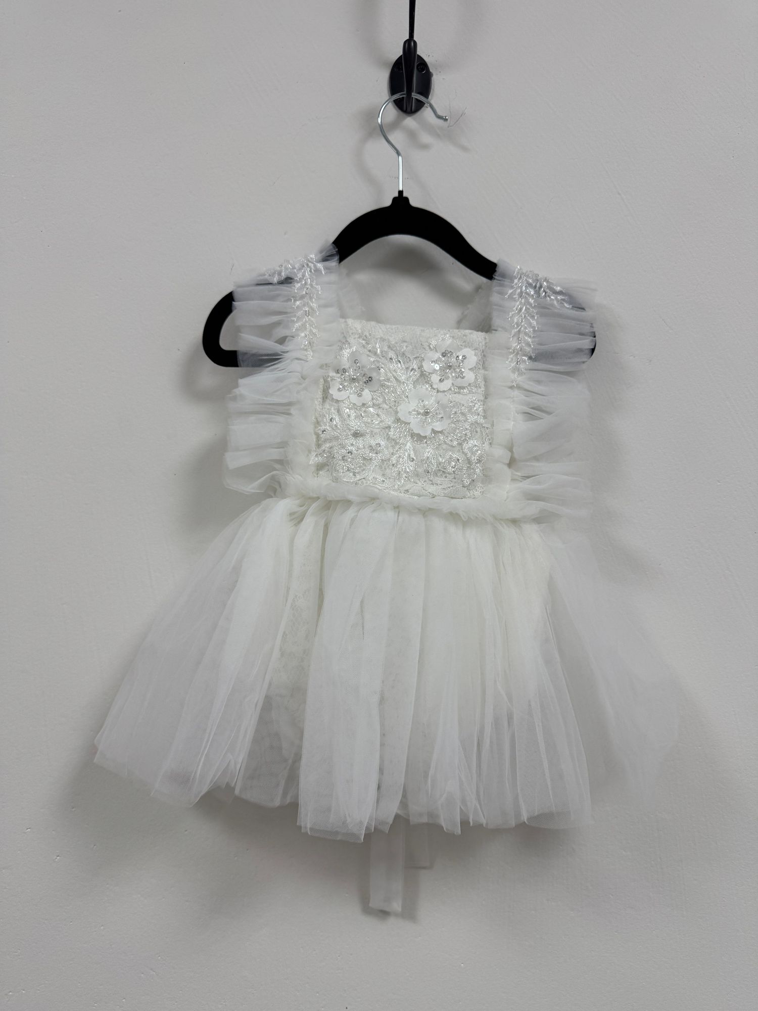 Delicate white tulle flower girl dress with lace bodice and ruffled cap sleeves hanging on black hanger against grey wall.