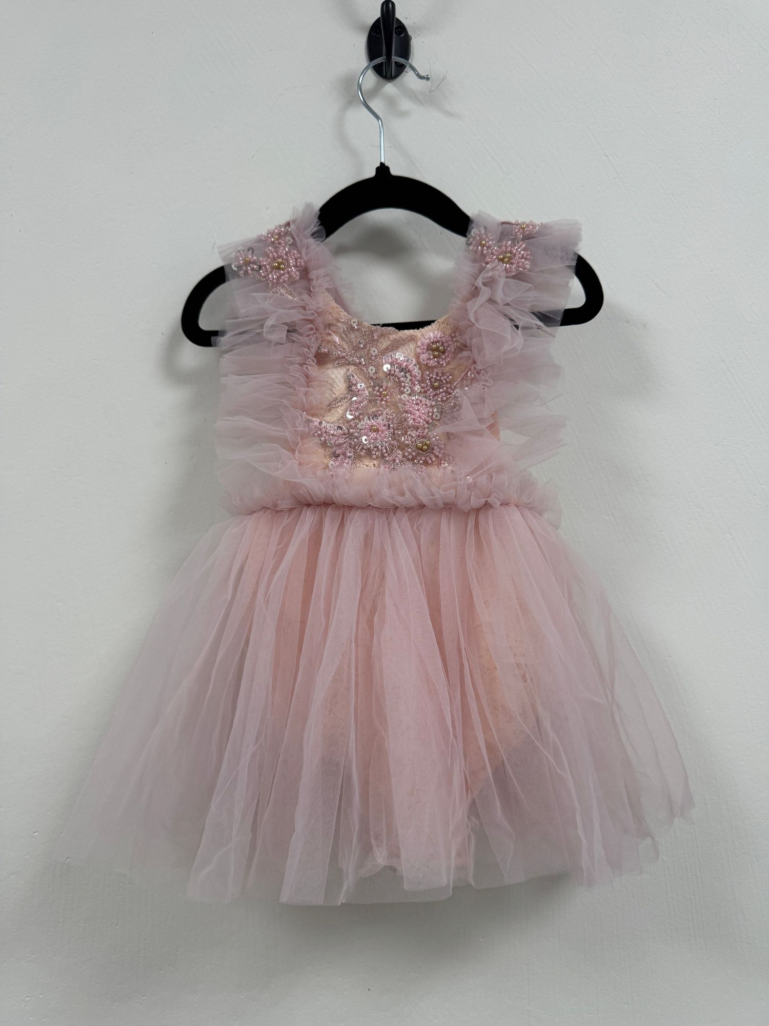 Delicate pink tulle dress with sequined bodice and ruffled straps hanging on black hanger against white wall.