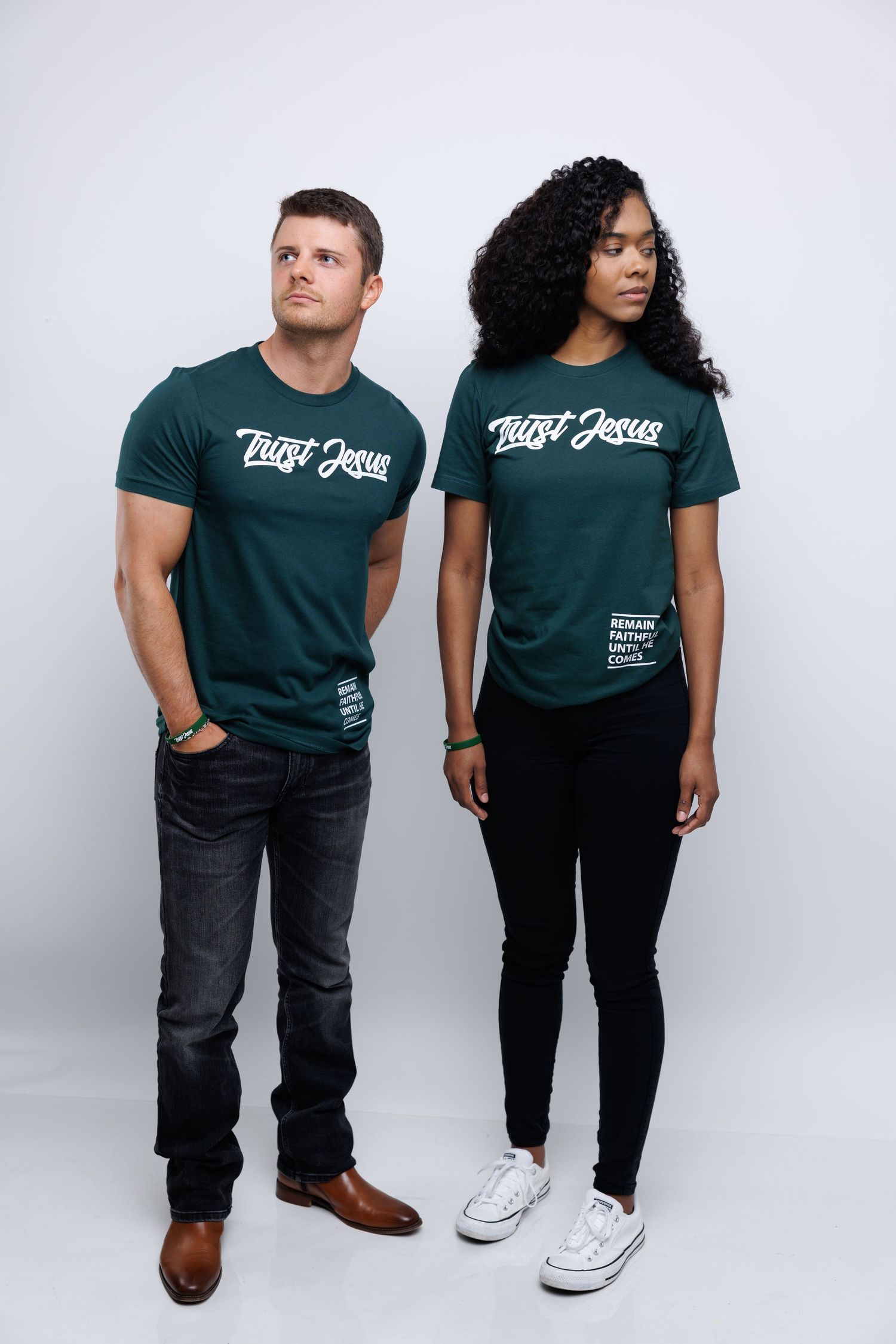 Two people stand together wearing matching dark green shirts with a logo against a gray background.
