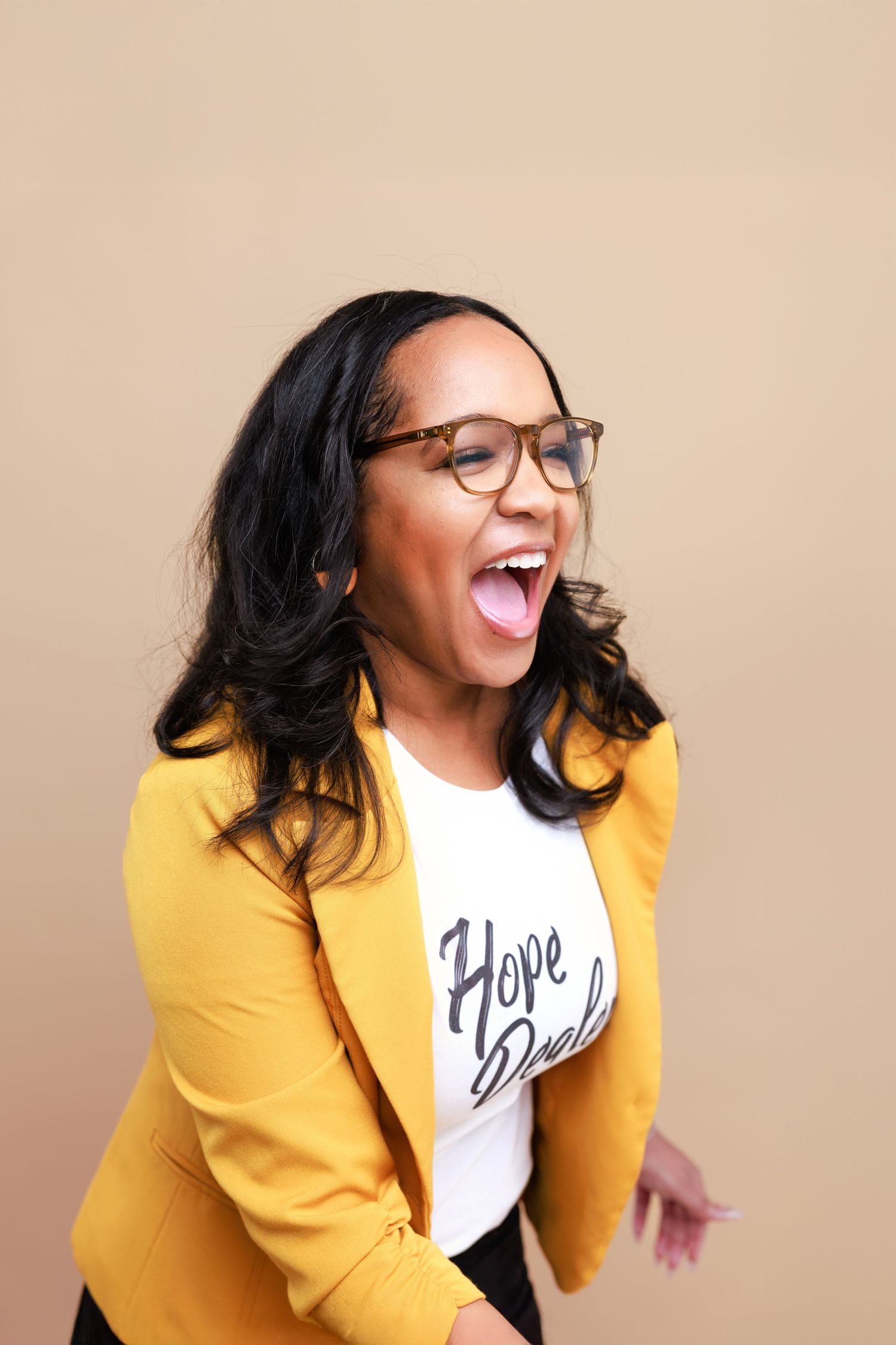 Person in yellow blazer and glasses laughing joyfully against beige background.