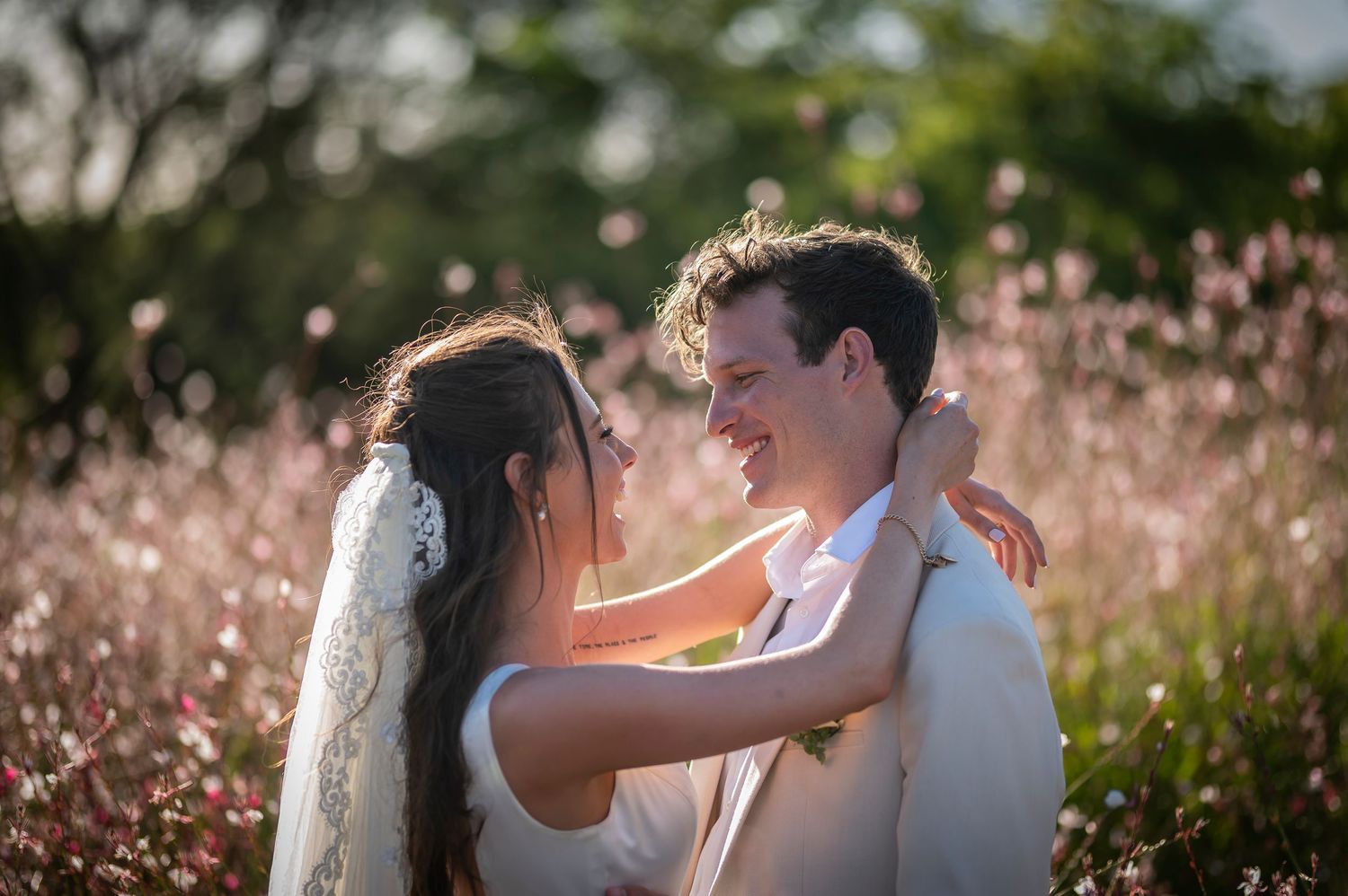 Newlywed couple embraces lovingly in a sun-lit meadow filled with pink wildflowers at golden hour.