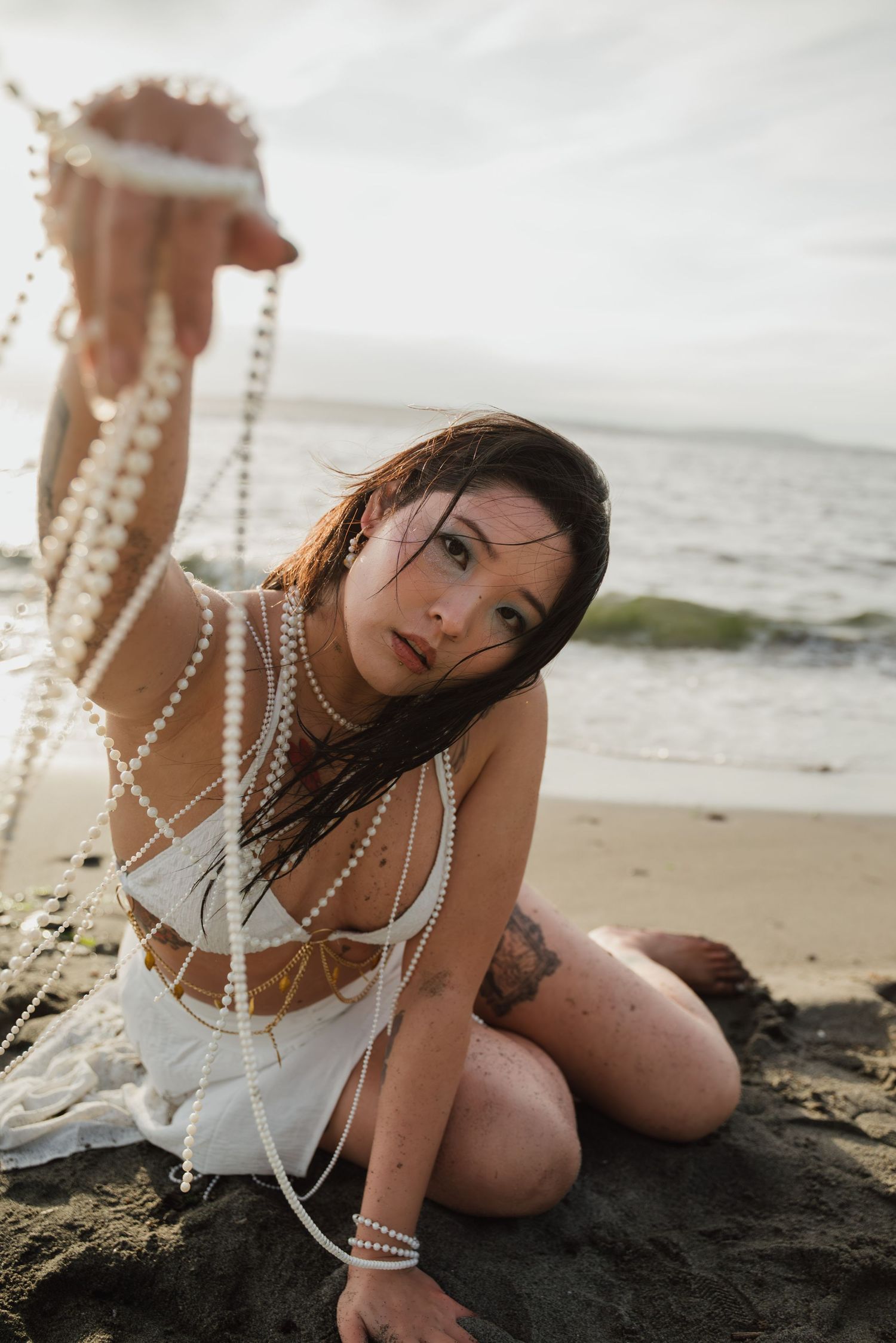 A beachwear fashion portrait series showing a model posing with pearl necklaces on black sand at sunset.