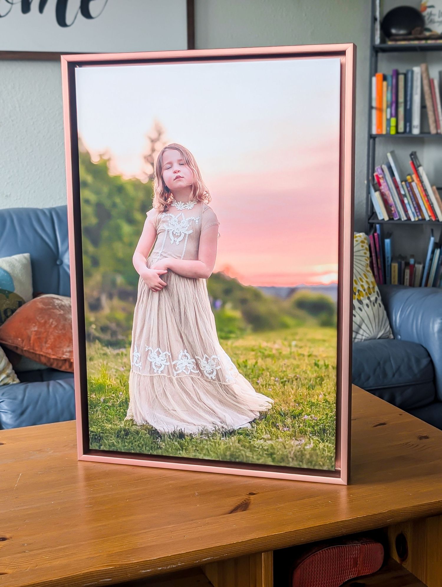 A framed portrait photograph in golden lighting displays a figure in a flowing cream dress standing on grassy hillside at sunset.