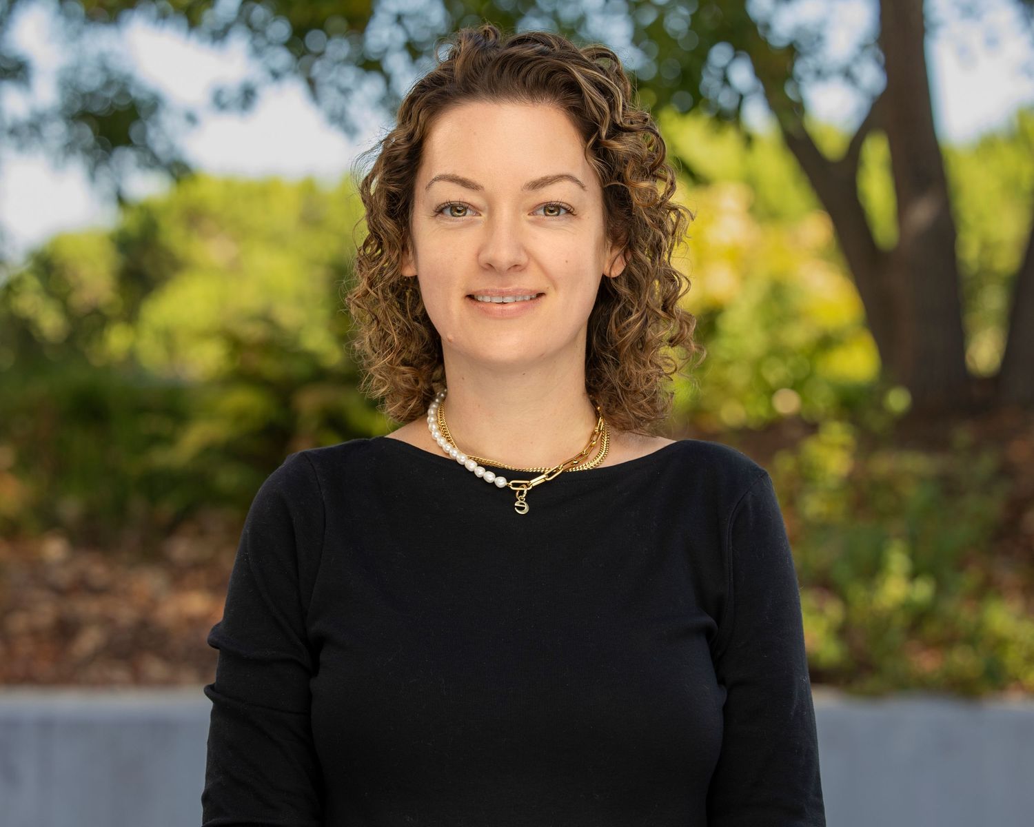 Professional headshot of smiling person wearing black sweater posed outdoors with greenery in background.