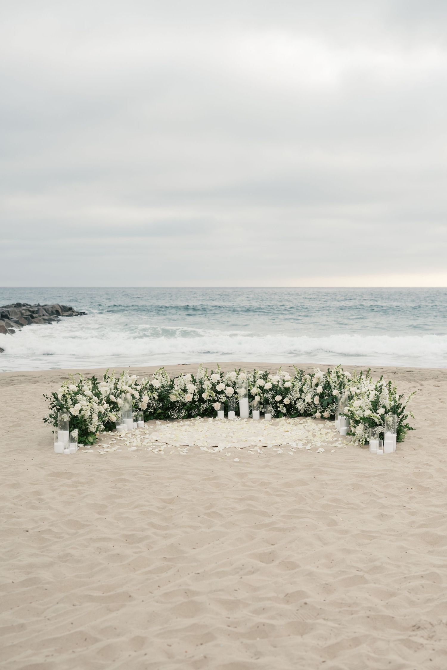 White flowers arranged in a heart shape on a sandy beach with ocean waves and cloudy skies in the background.